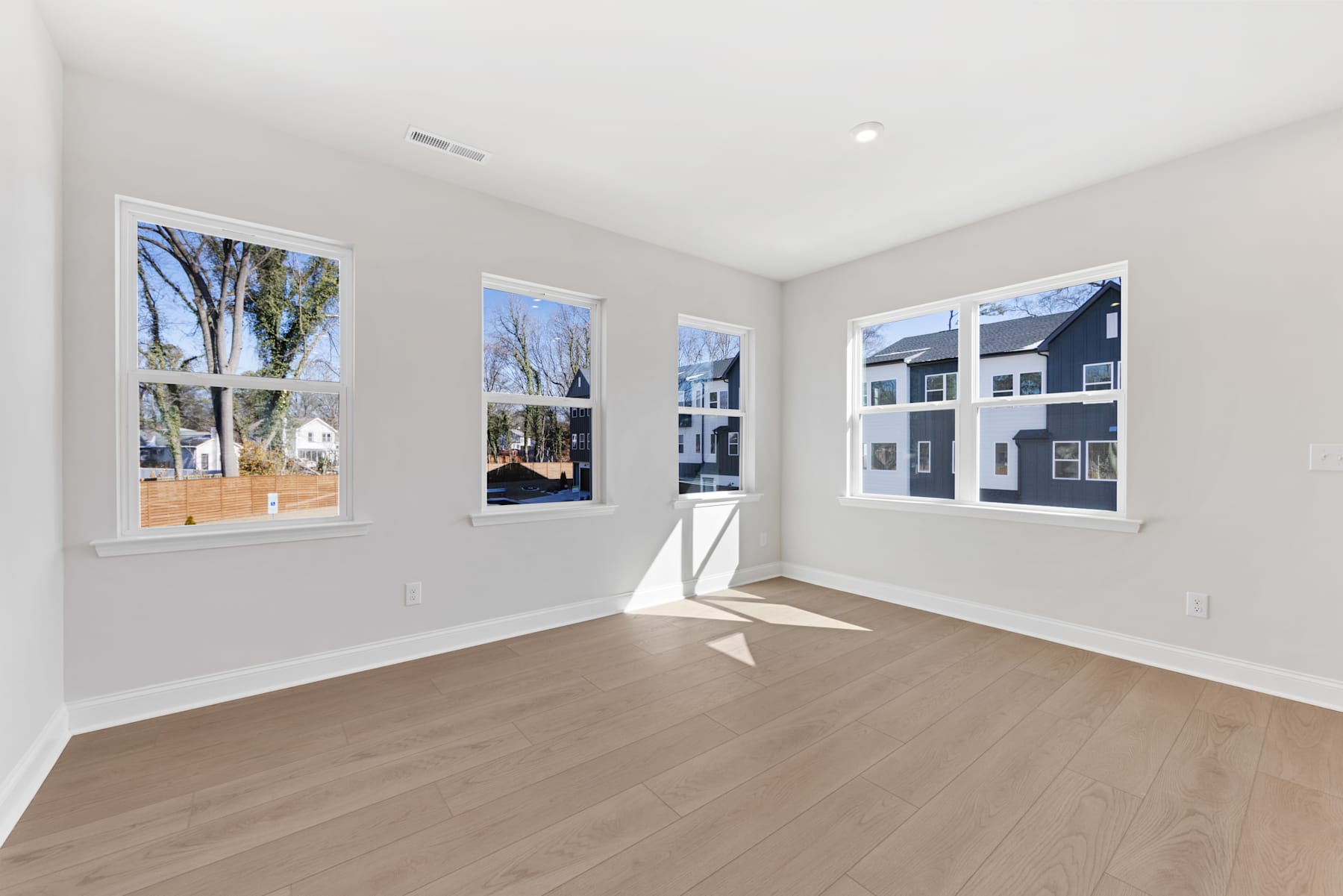 A bright and spacious room with large windows overlooking a residential neighborhood, featuring hardwood floors and neutral-colored walls.