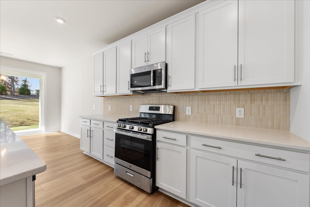 A modern, well-equipped kitchen with white cabinets, stainless steel appliances, and a tiled backsplash, set against a backdrop of hardwood floors and a window overlooking a grassy outdoor area.