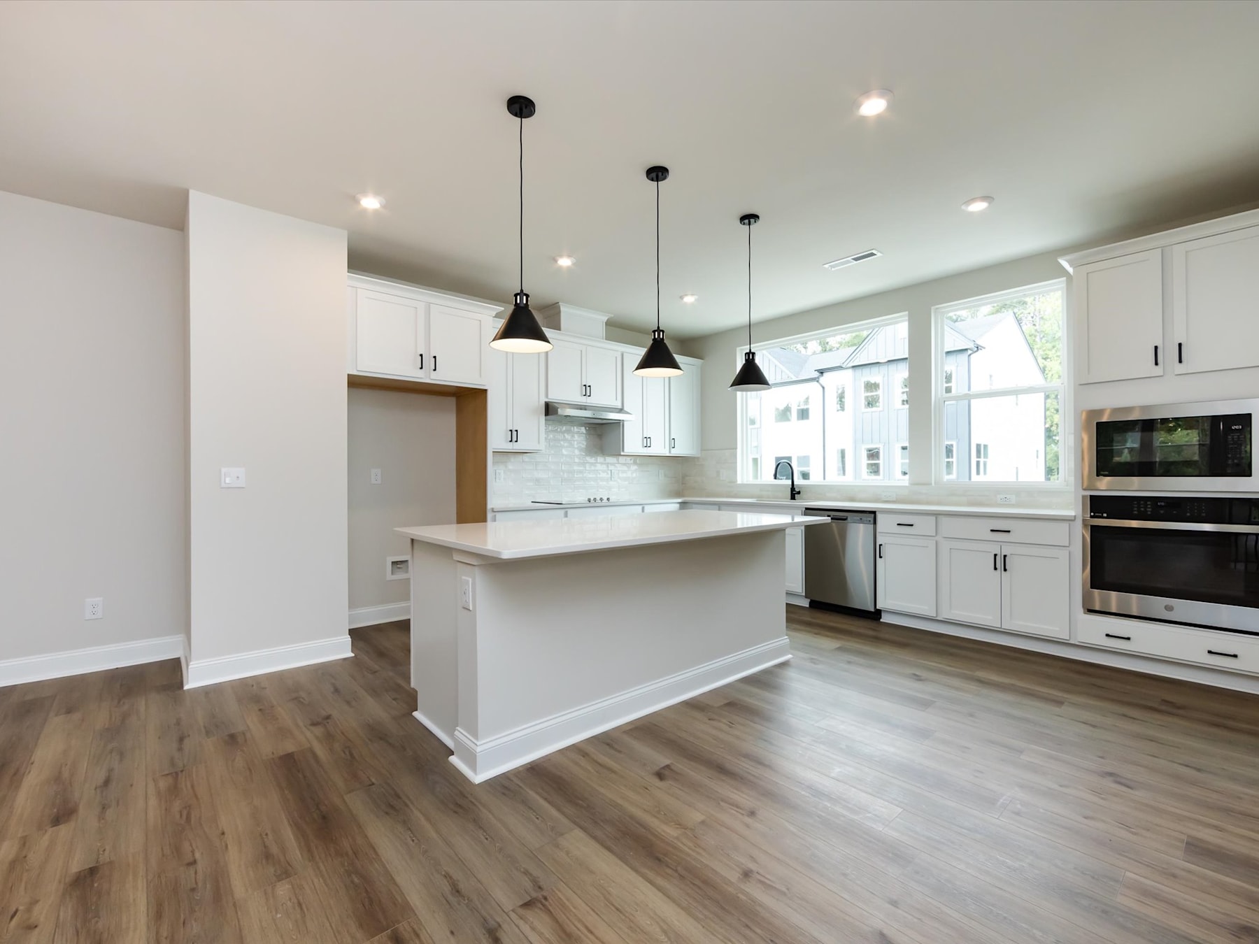 A modern, open-concept kitchen with white cabinets, a central island, and pendant lighting fixtures, set against a backdrop of hardwood floors and large windows.