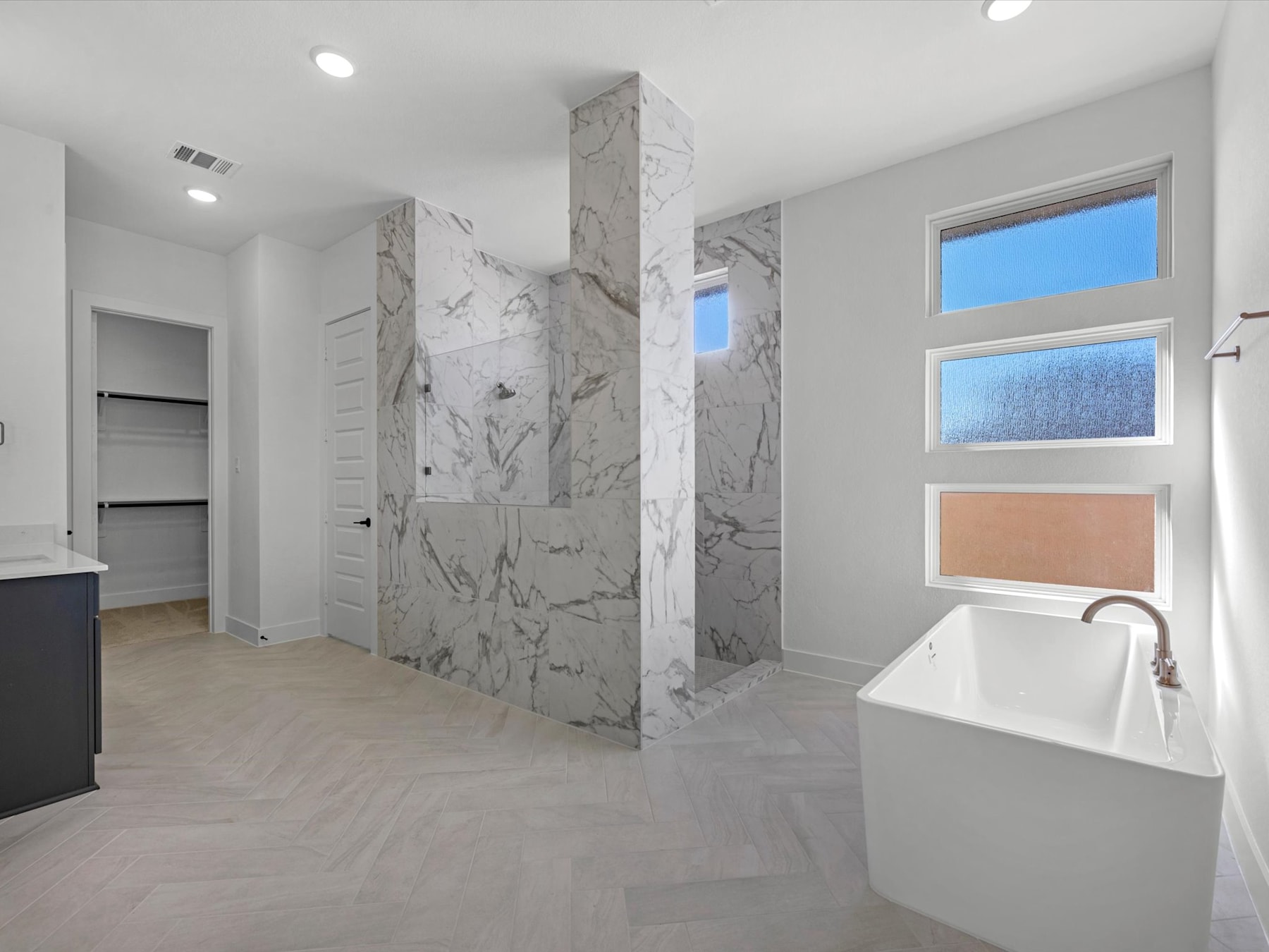 A modern, spacious bathroom with a freestanding bathtub, marble-like tiles covering the walls, and a large window providing natural light.