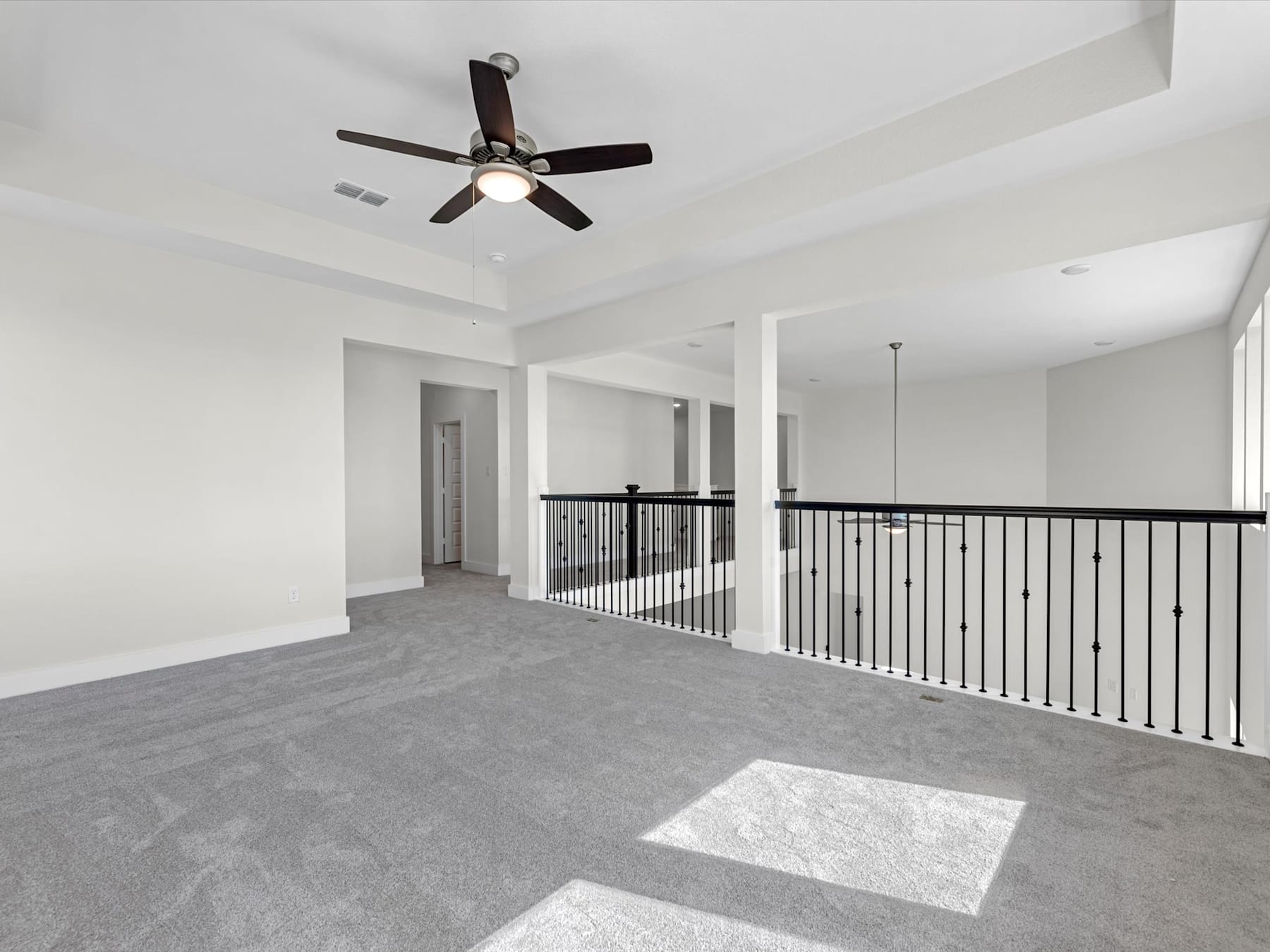 An empty, spacious room with a ceiling fan and a carpeted floor, leading to a hallway with a railing.