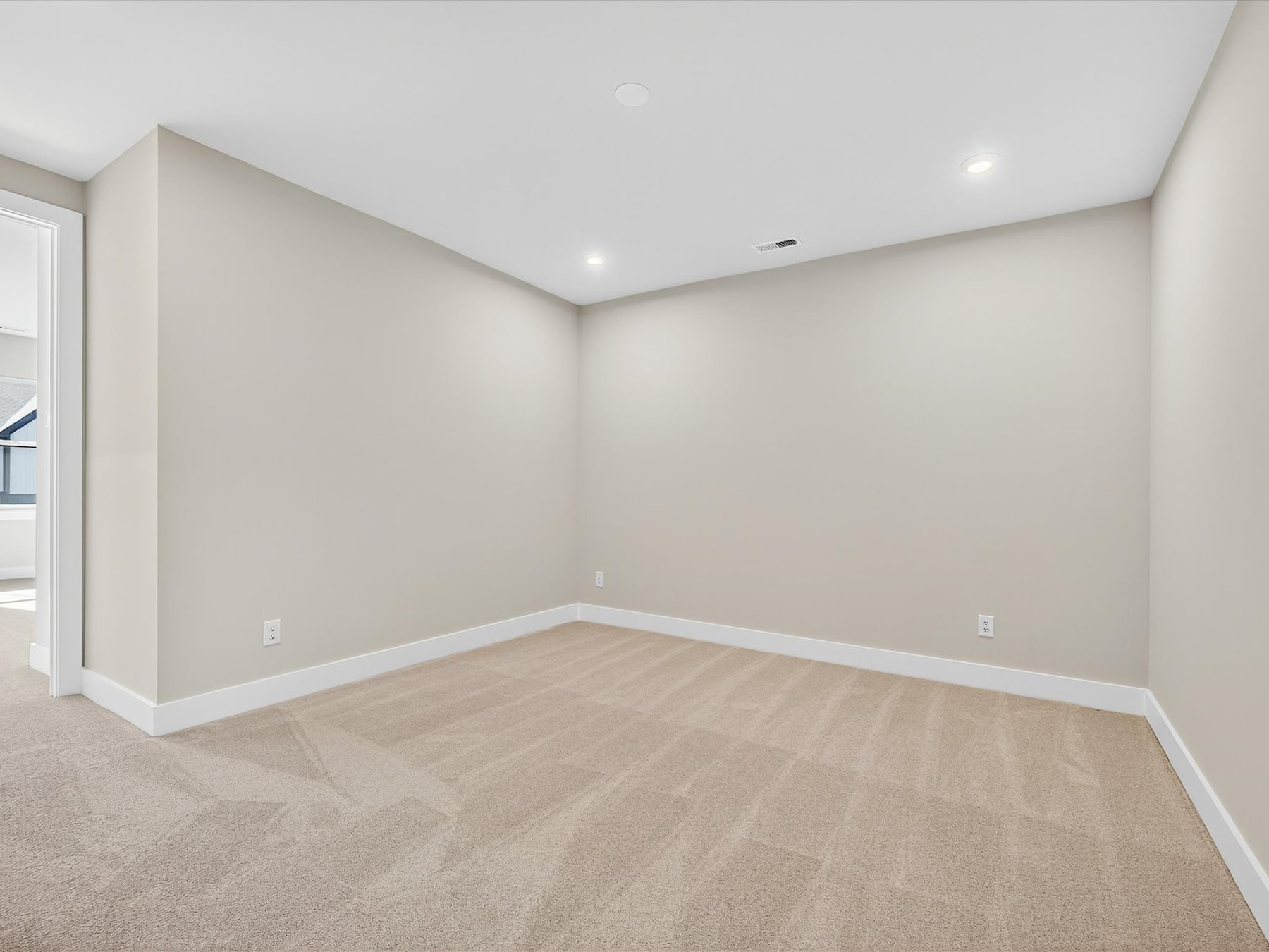 An empty room with beige walls and a light-colored hardwood floor, with a window visible in the background.