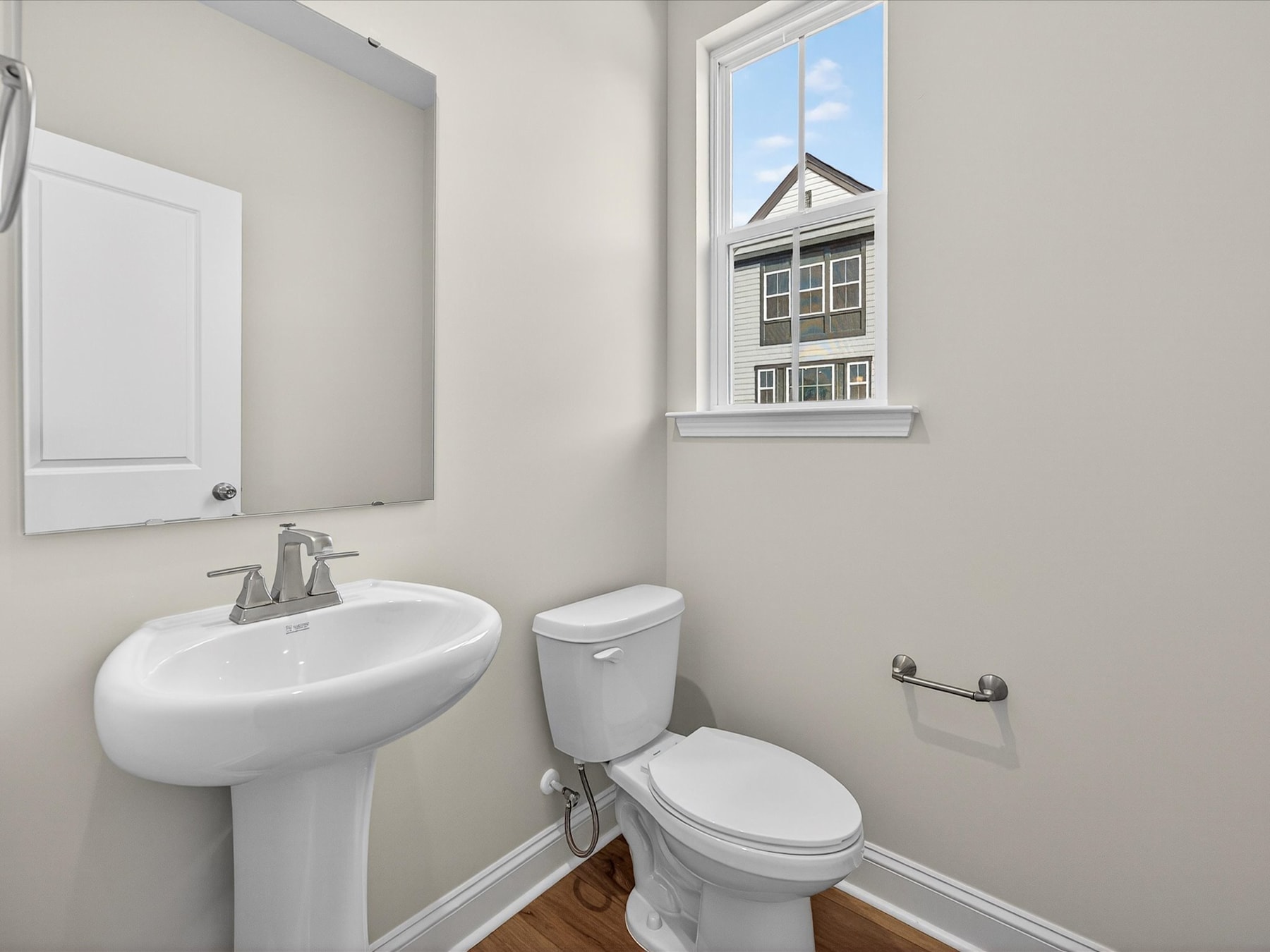 A clean, well-lit bathroom with a pedestal sink, toilet, and a window overlooking a residential building in the background.