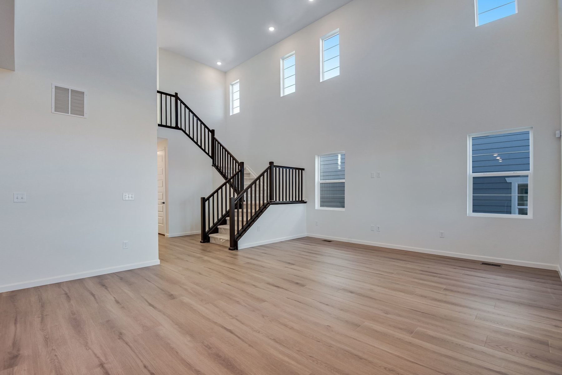 A spacious, well-lit room with a wooden floor, a staircase with black railings, and large windows allowing natural light to flood the space.