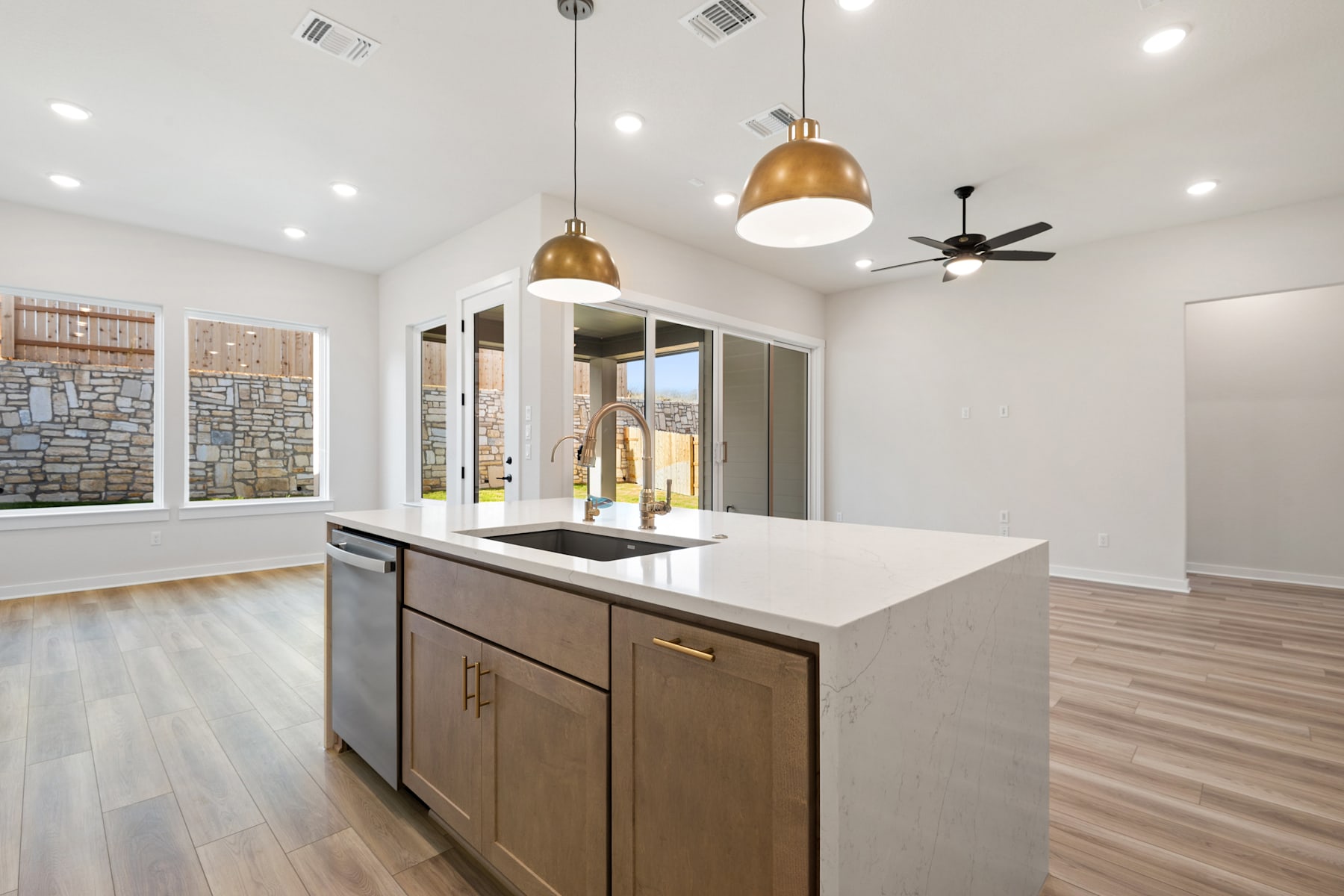 A modern and spacious kitchen with a central island, wooden cabinets, and pendant lighting fixtures, set against a backdrop of large windows and a stone wall.