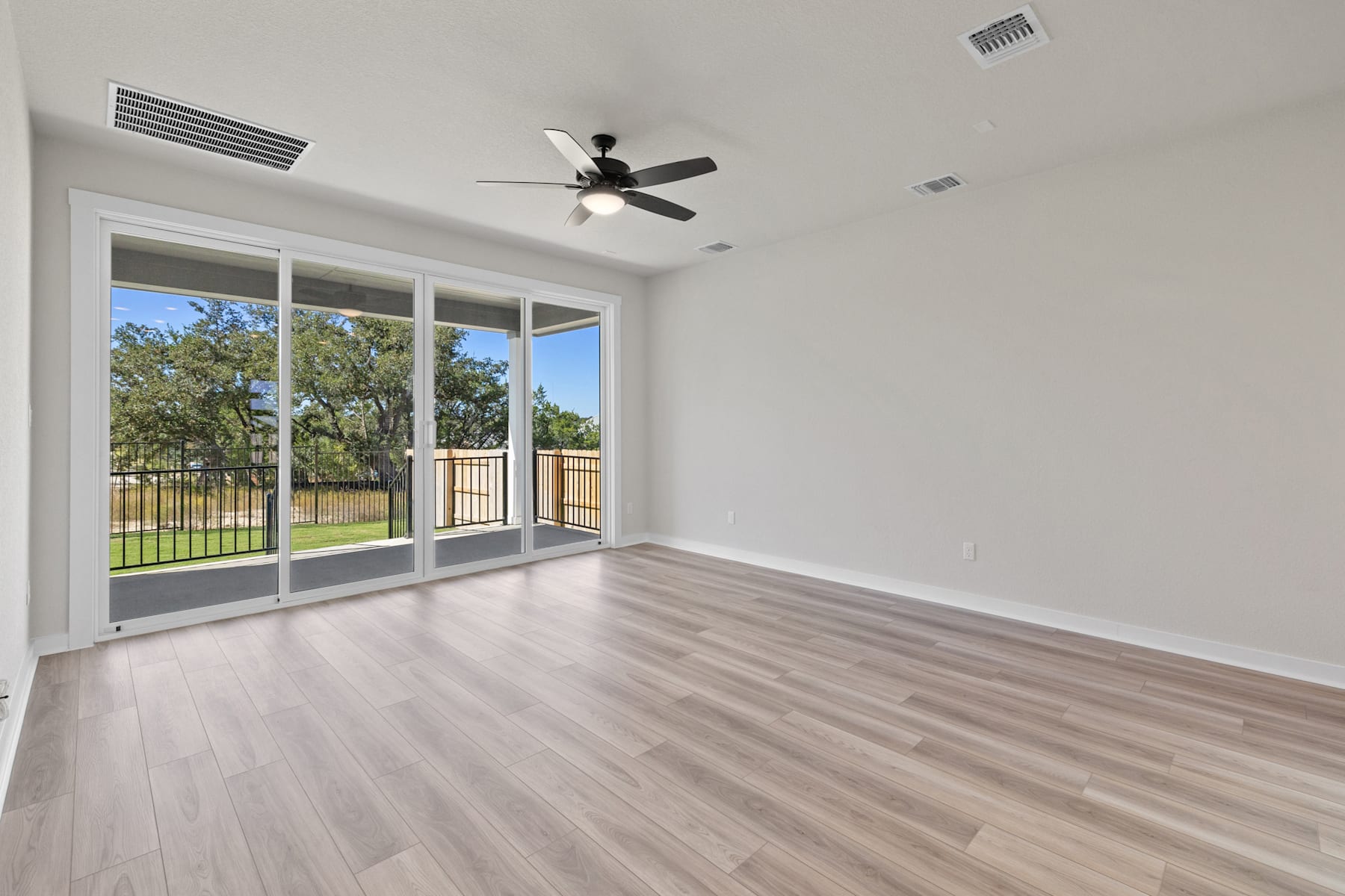 A spacious, well-lit room with a ceiling fan, large windows overlooking a fenced outdoor area, and a hardwood floor.