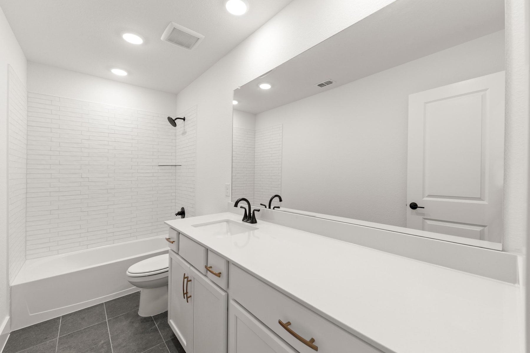 A modern, minimalist bathroom with white walls, a vanity with drawers, and a large mirror above it, as well as a toilet and a shower visible in the background.