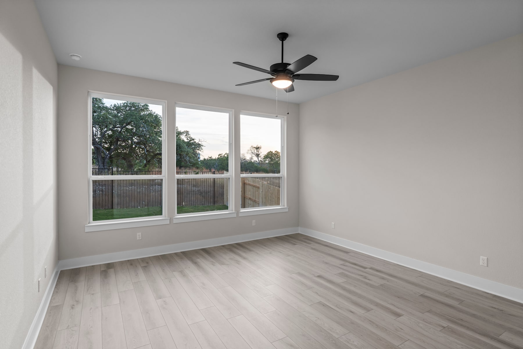 A bright and airy room with a ceiling fan, large windows overlooking a backyard, and a hardwood floor.