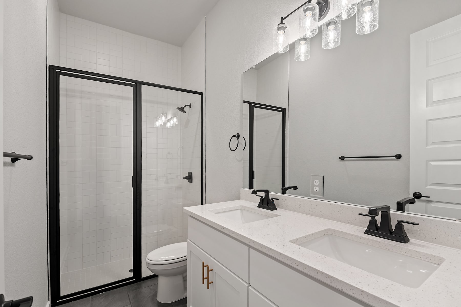 A modern, minimalist bathroom with a white vanity, black framed mirrors, and a glass shower enclosure, illuminated by a stylish chandelier.