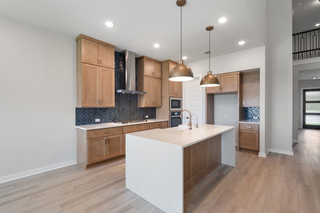 A modern and spacious kitchen with light wood cabinets, a white island, and pendant lighting fixtures, set against a backdrop of hardwood floors and white walls.