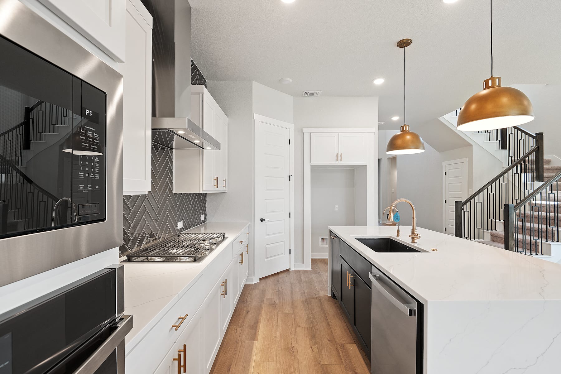 A modern, open-concept kitchen with white cabinets, stainless steel appliances, and warm brass lighting fixtures, set against a backdrop of a hallway and staircase.