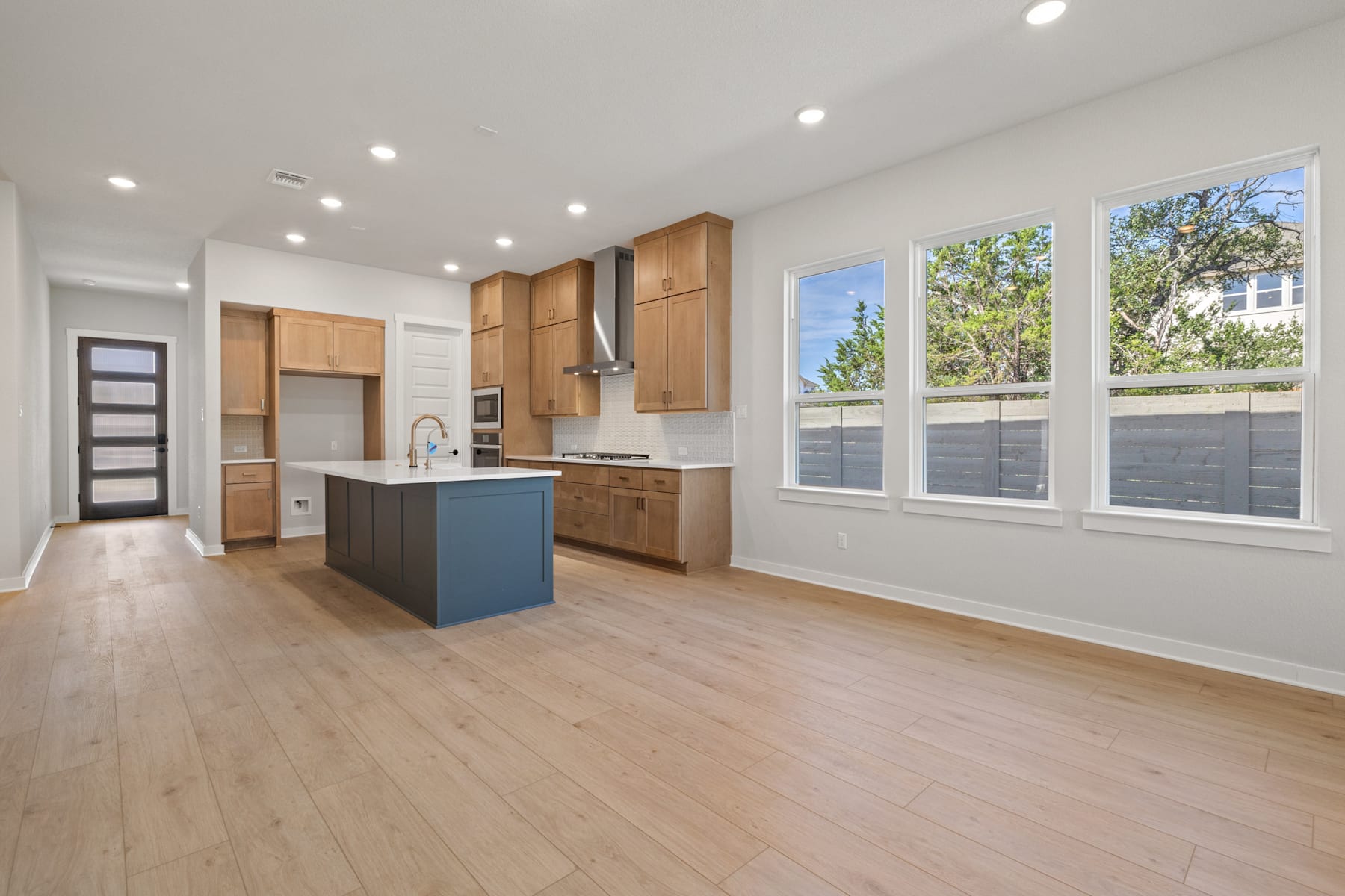 A modern, open-concept kitchen with wooden cabinets, a blue island, and large windows overlooking a lush outdoor area.