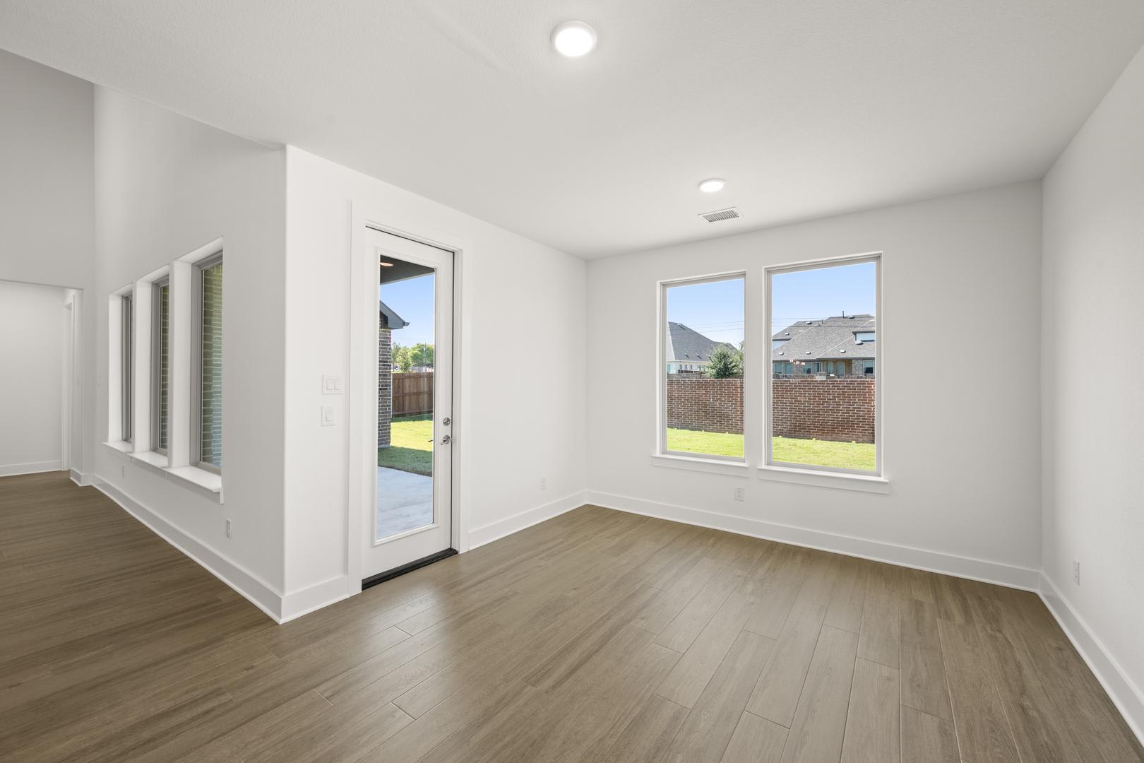 A spacious, well-lit room with white walls, hardwood floors, and large windows overlooking a backyard.