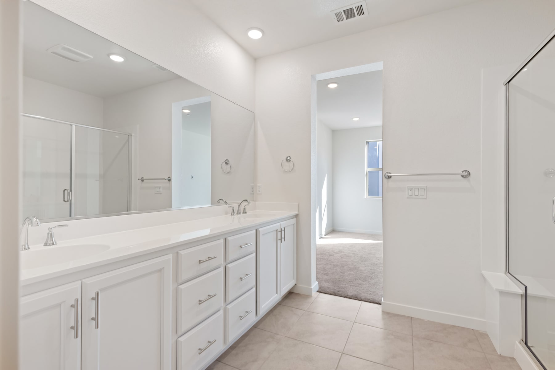 A spacious and well-lit bathroom with a large vanity, multiple drawers, and a mirror extending across the wall. The floor is covered in light-colored tiles, and the room appears to be part of a larger, modern living space.