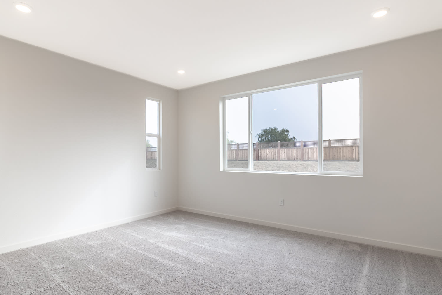 A bright, empty room with large windows overlooking a backyard area.
