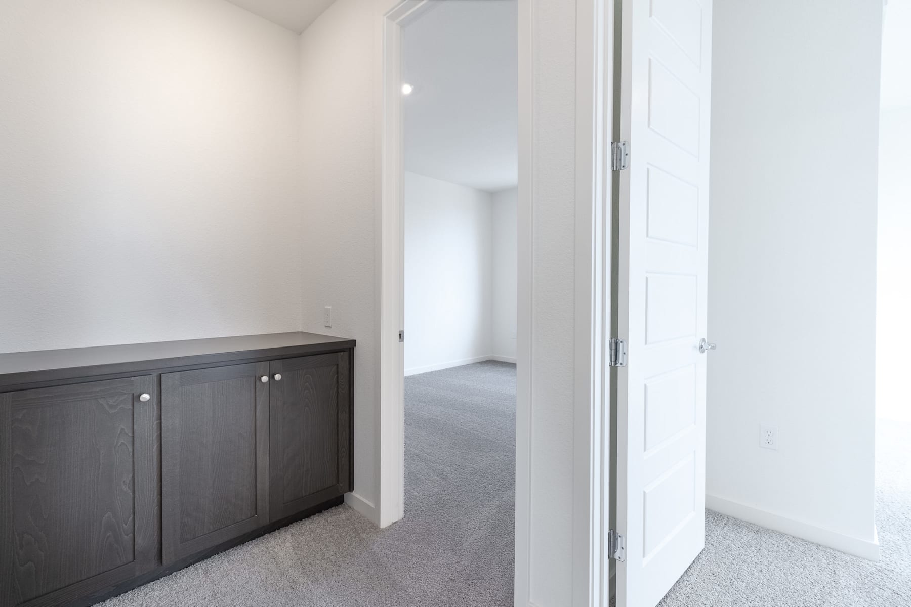 A dark wooden cabinet stands in the foreground of a bright, minimalist room with white walls and a partially visible doorway in the background.