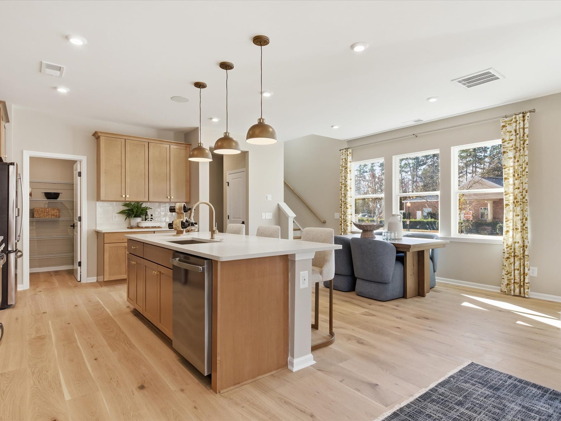 A modern, open-concept kitchen with light wood cabinets, a central island, and large windows overlooking a cozy living area.