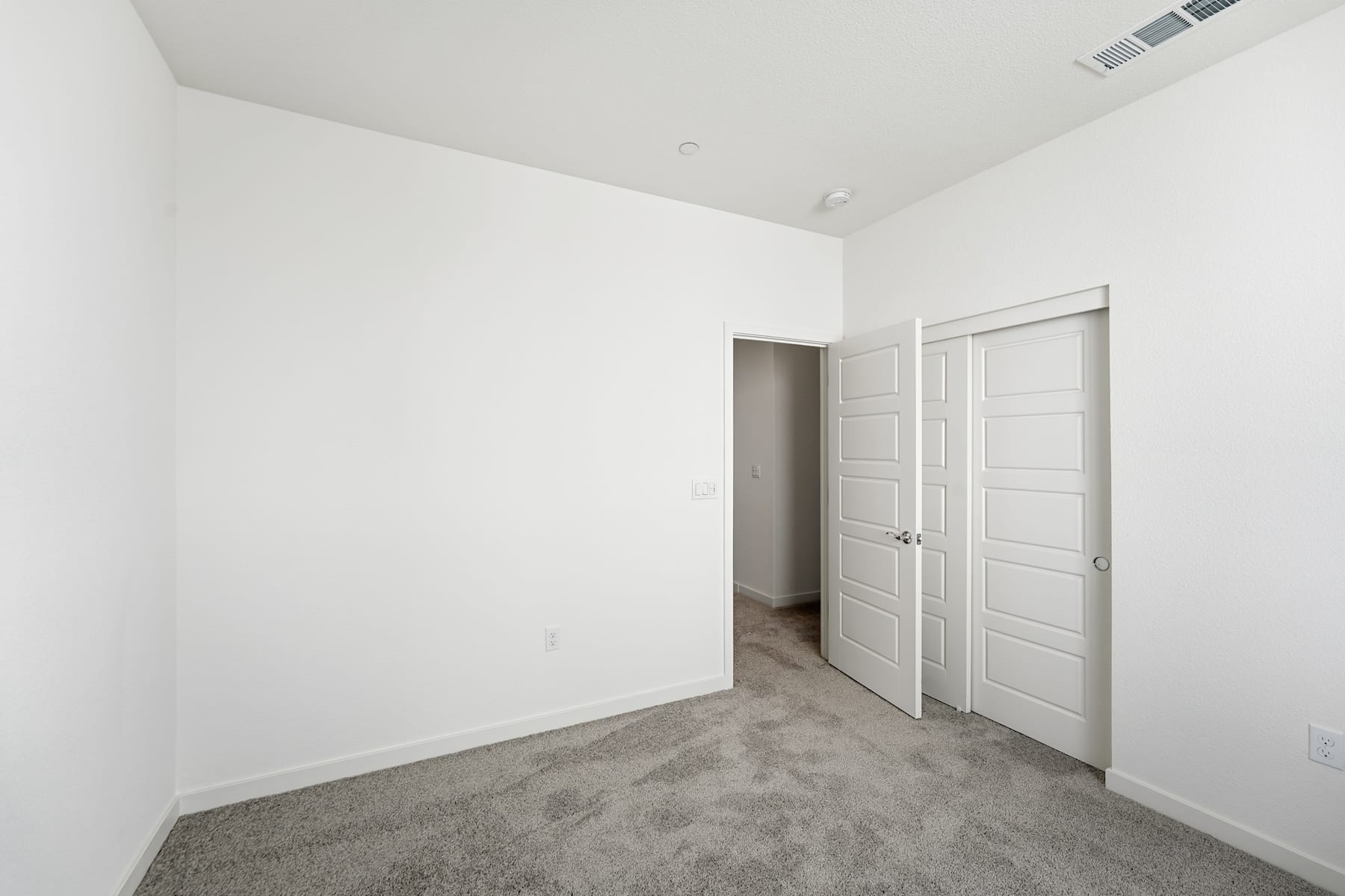 A simple, minimalist bedroom with white walls, a gray carpeted floor, and two closed doors leading to other rooms.