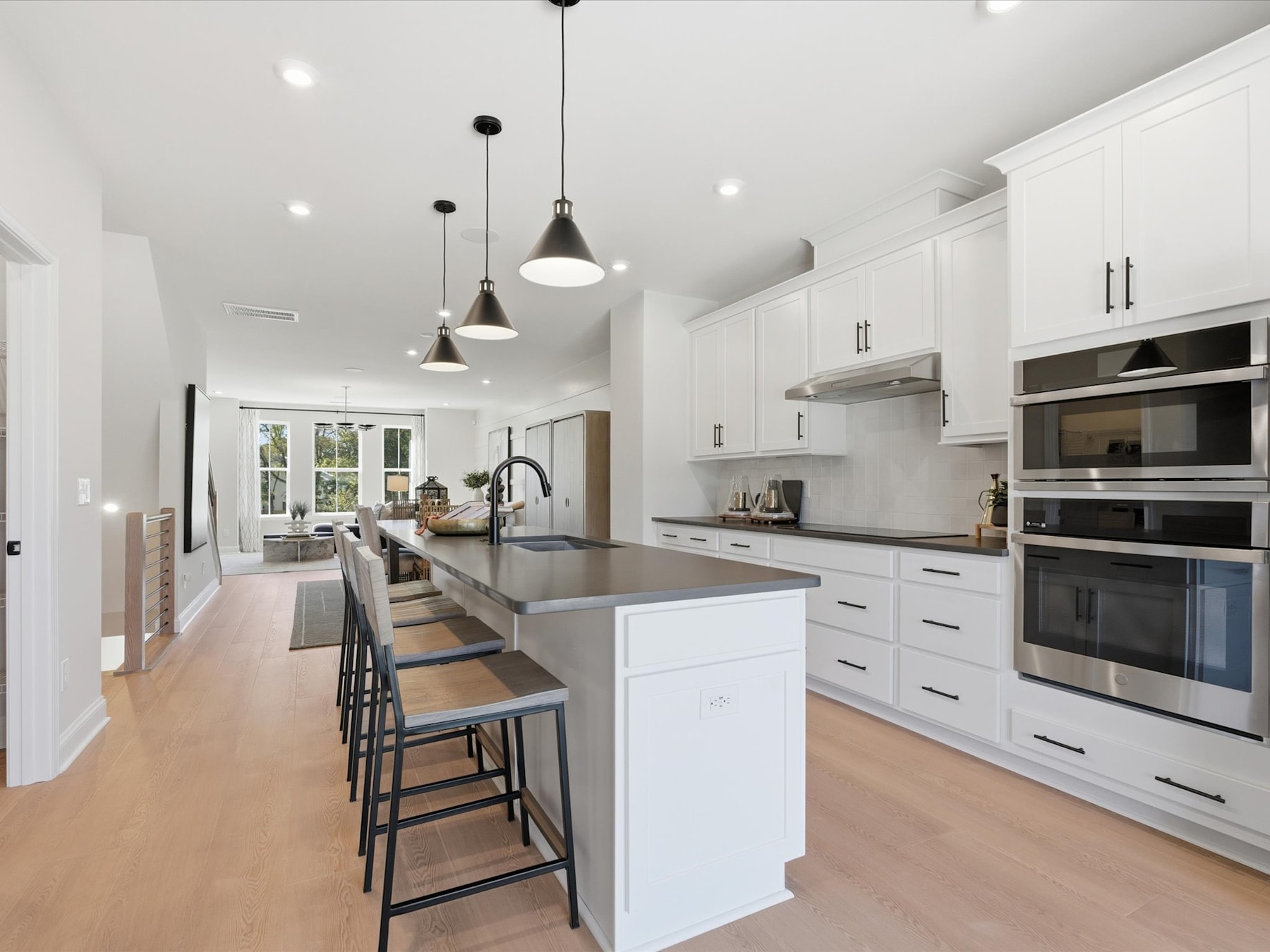 A modern, open-concept kitchen with white cabinets, stainless steel appliances, and a central island with bar stools, set against a backdrop of large windows and natural light.