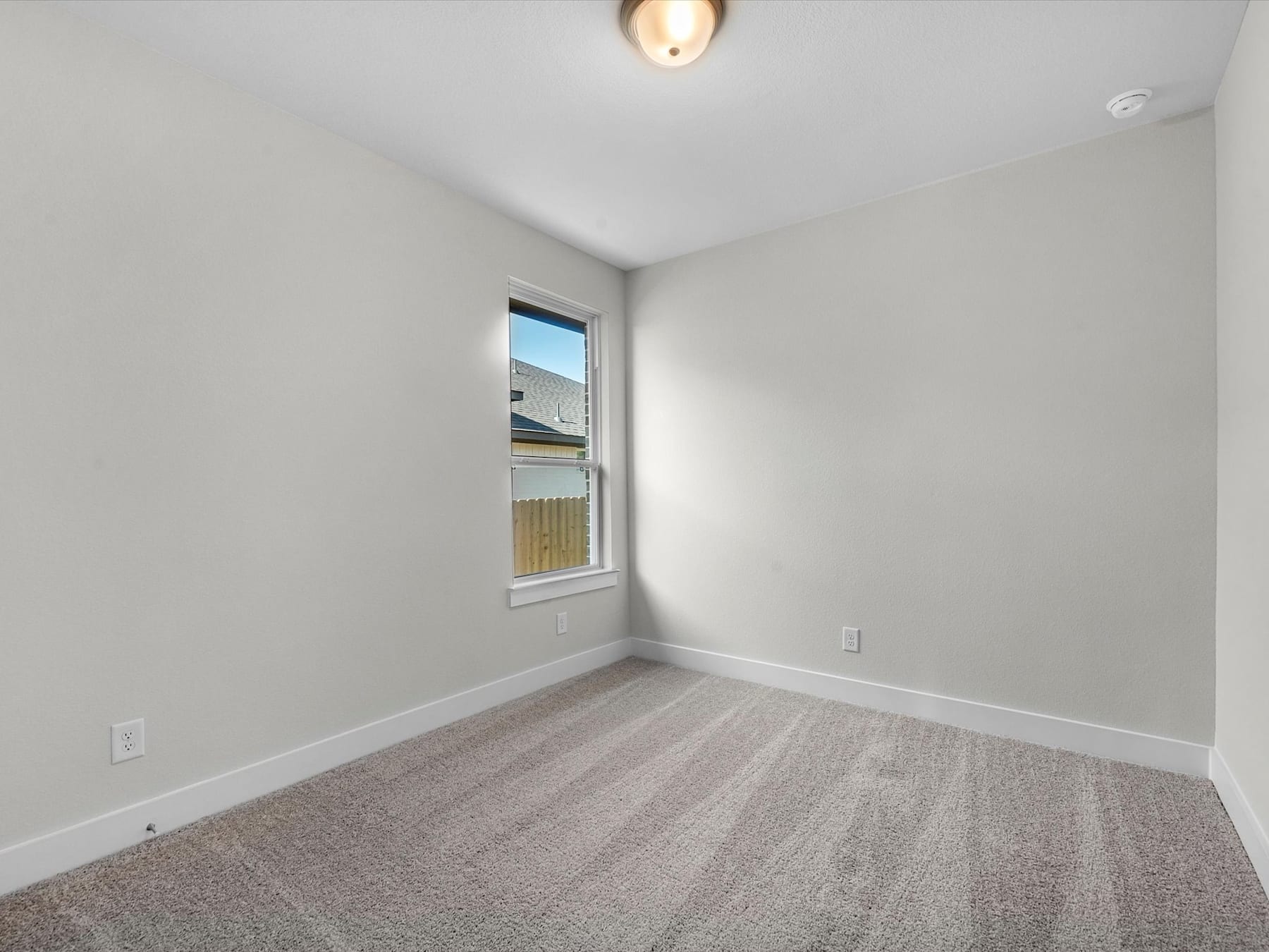 A sparsely furnished room with a window, a light fixture, and a plain gray carpet covering the floor.