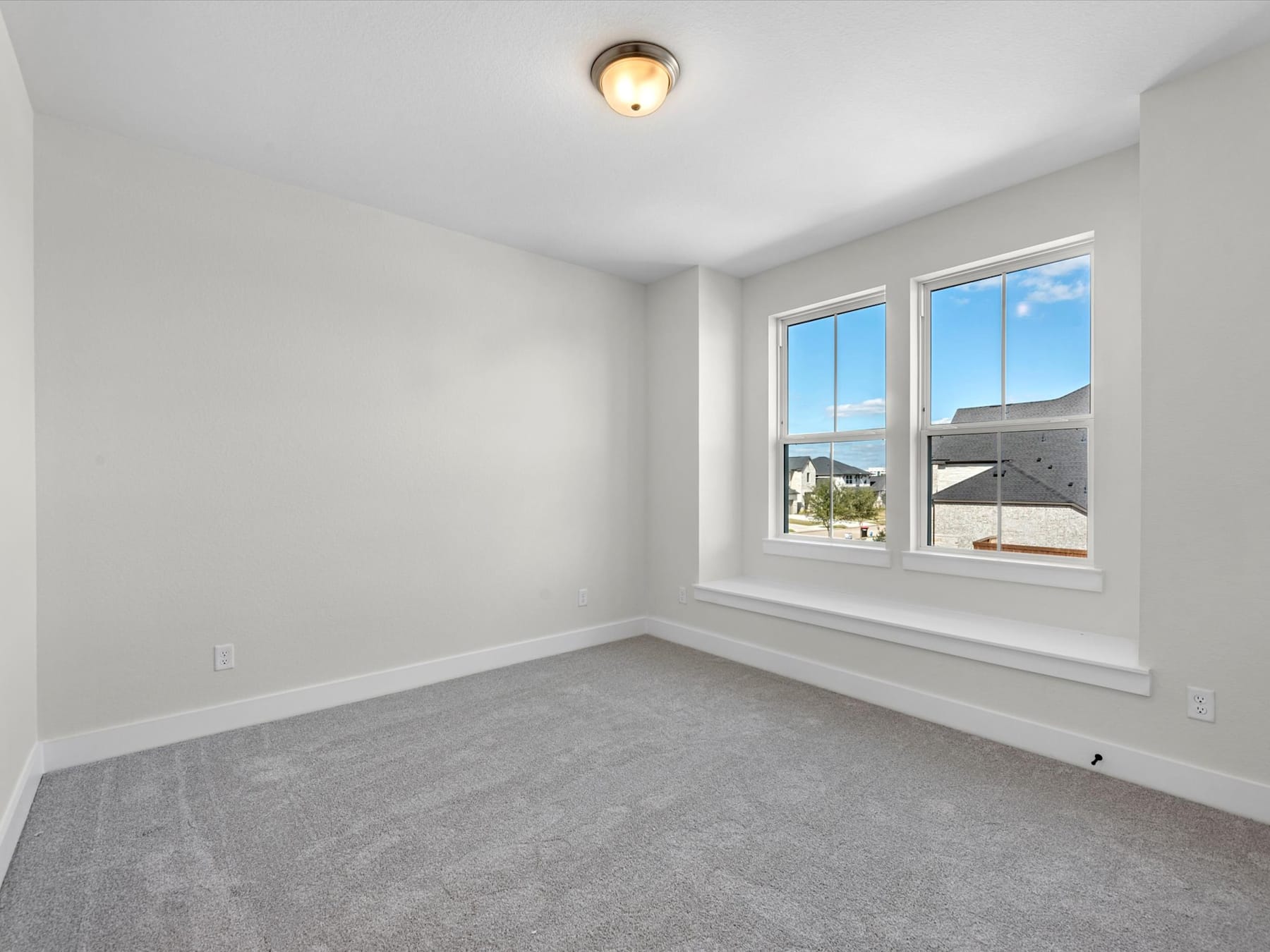 A bright, empty room with a large window overlooking a residential neighborhood, featuring a simple light fixture and gray carpeted floor.