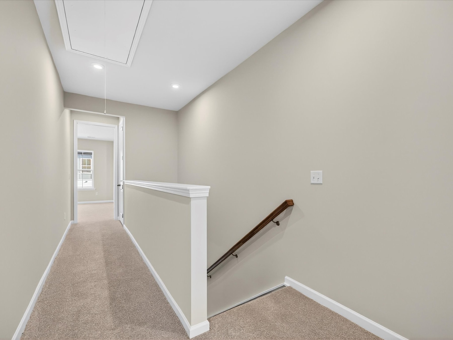 A hallway with a carpeted floor, white walls, and a wooden handrail leading to an open doorway.