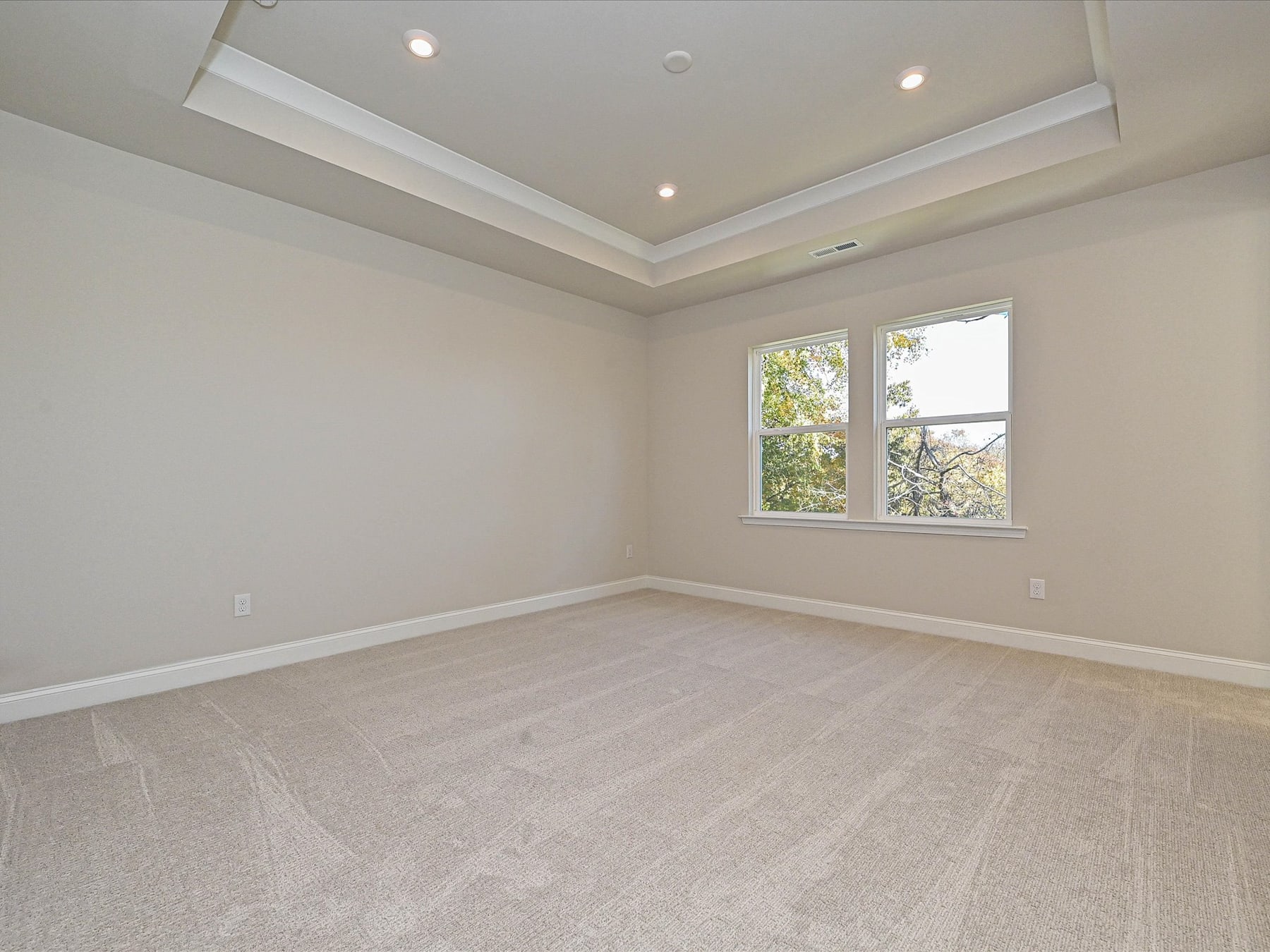 A spacious, well-lit room with a tray ceiling, hardwood floors, and large windows overlooking the outdoors.