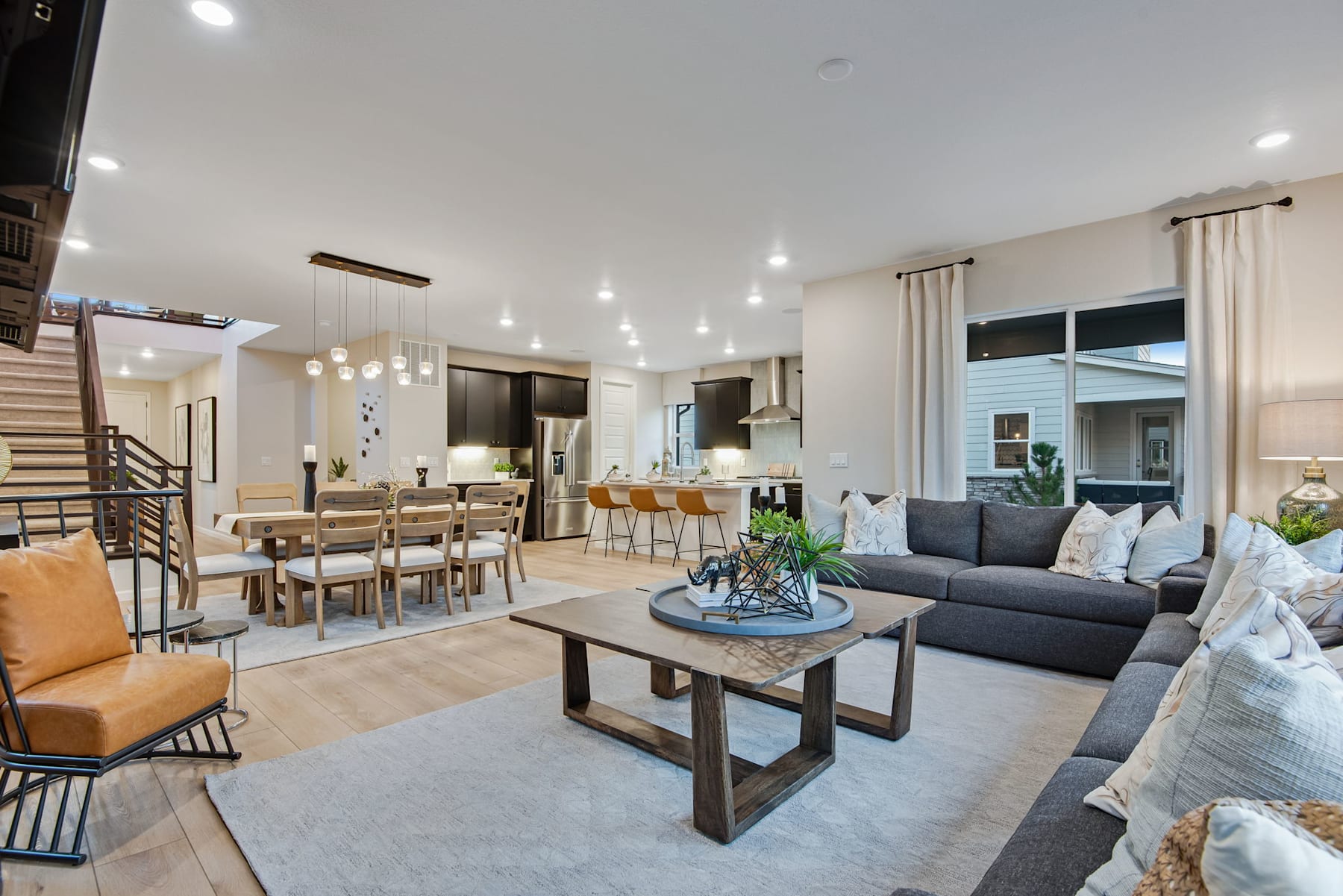 A spacious and modern open-concept living room with a dining area and kitchen, featuring a gray sectional sofa, a wooden coffee table, and various lighting fixtures creating a warm and inviting atmosphere.