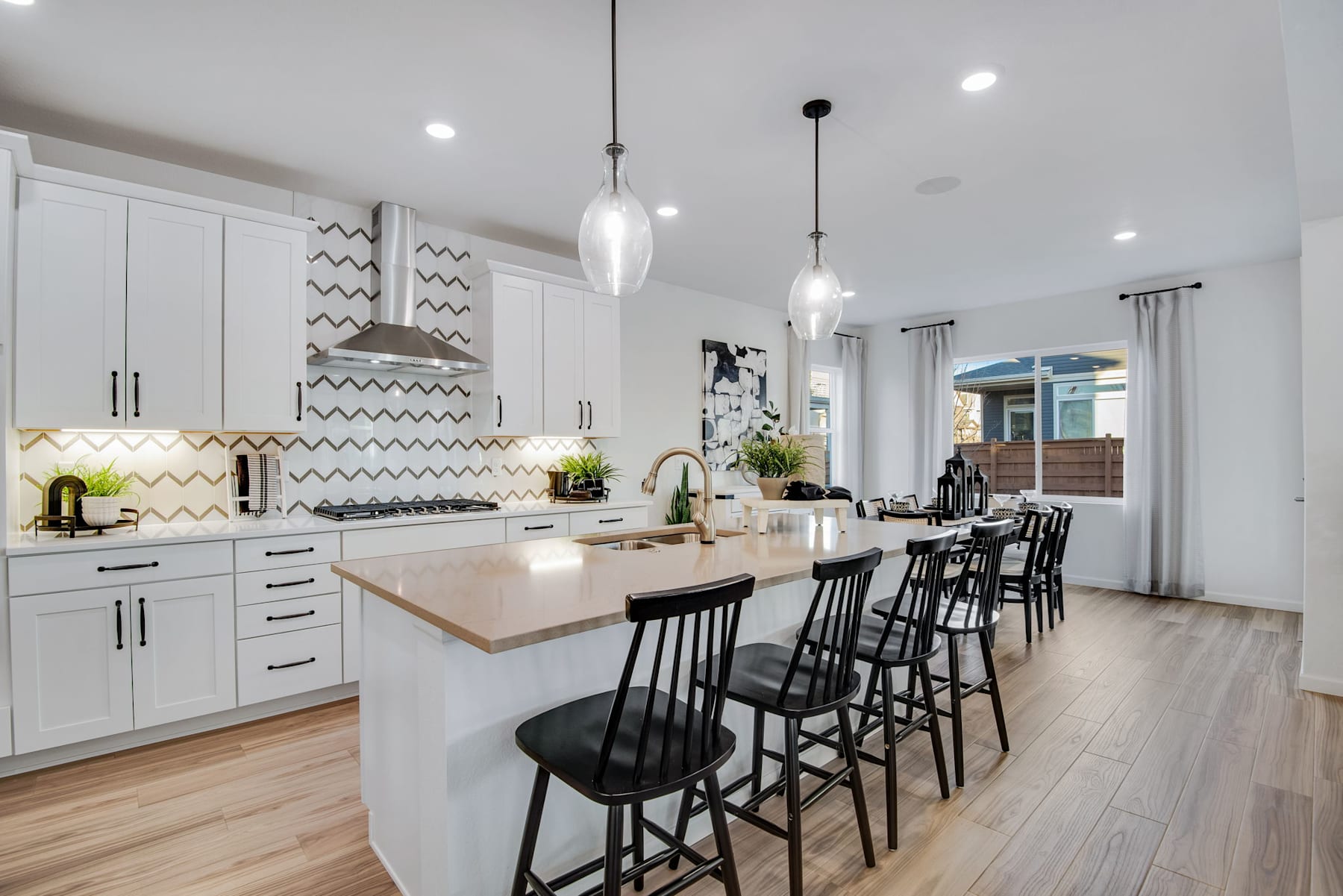 A modern, open-concept kitchen with white cabinets, a large island with seating, and pendant lighting fixtures, set against a backdrop of hardwood floors and a sliding glass door leading to an outdoor area.