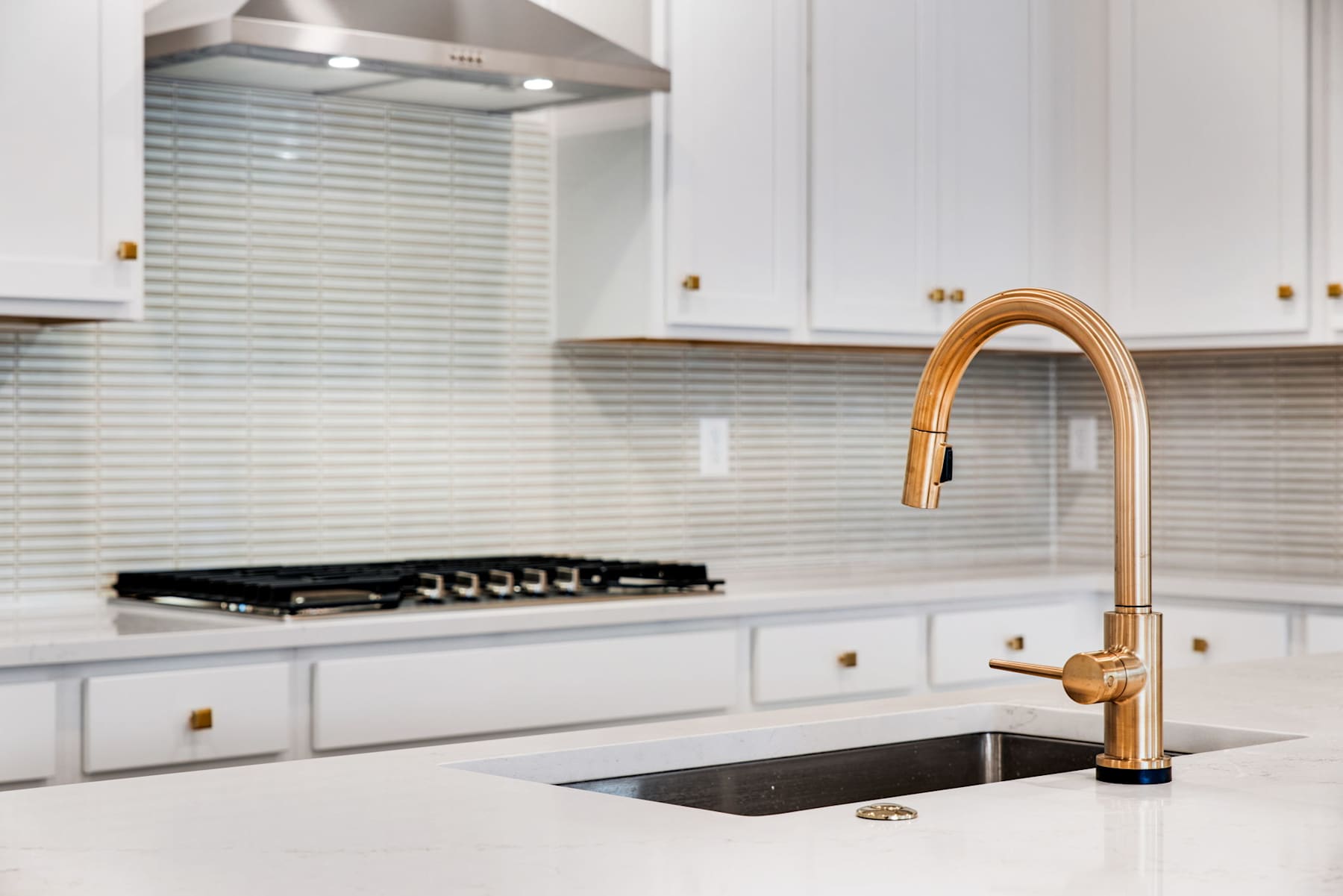 A modern kitchen with white cabinets, a stainless steel range hood, and a gold-colored faucet over a black sink.