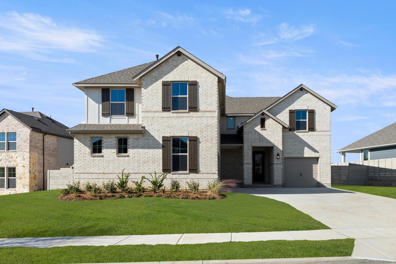 A two-story residential house with a well-manicured lawn and landscaping in the foreground, set against a clear blue sky with scattered clouds in the background.