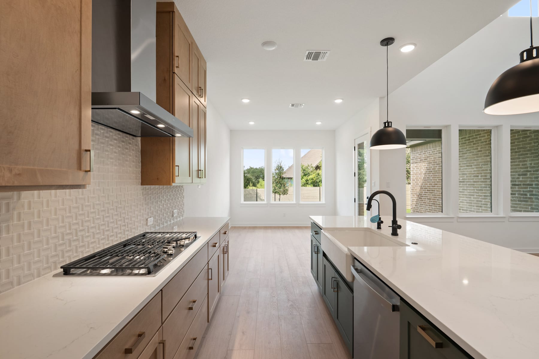A modern, well-lit kitchen with wooden cabinets, a white countertop, and a tiled backsplash, featuring a gas stove and a window overlooking the exterior.