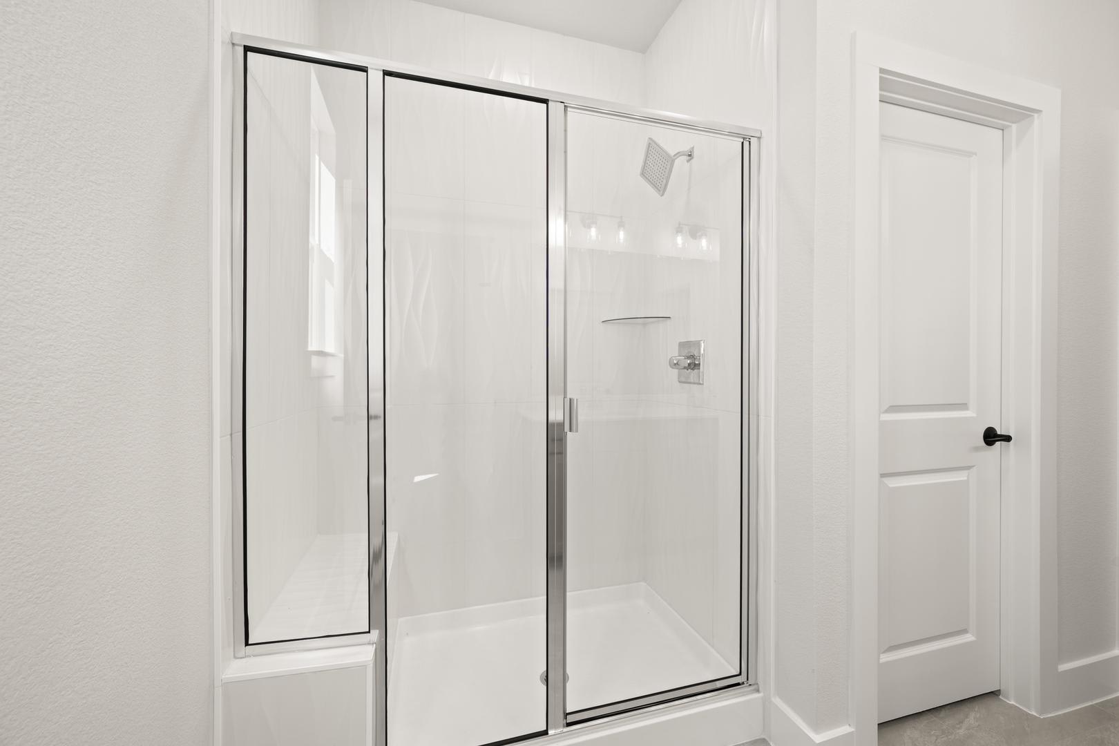 A white, minimalist bathroom with a glass-enclosed shower stall and a closed door in the background.