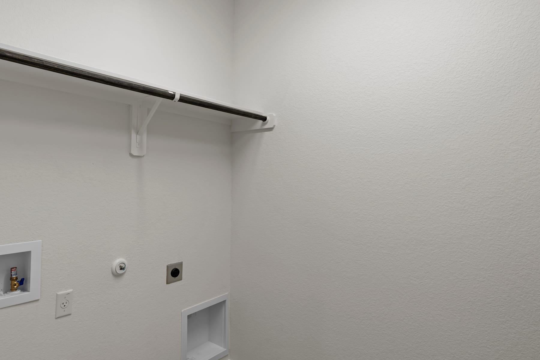 A simple, minimalist closet space with a white wall, a metal clothing rod, and an electrical outlet visible.