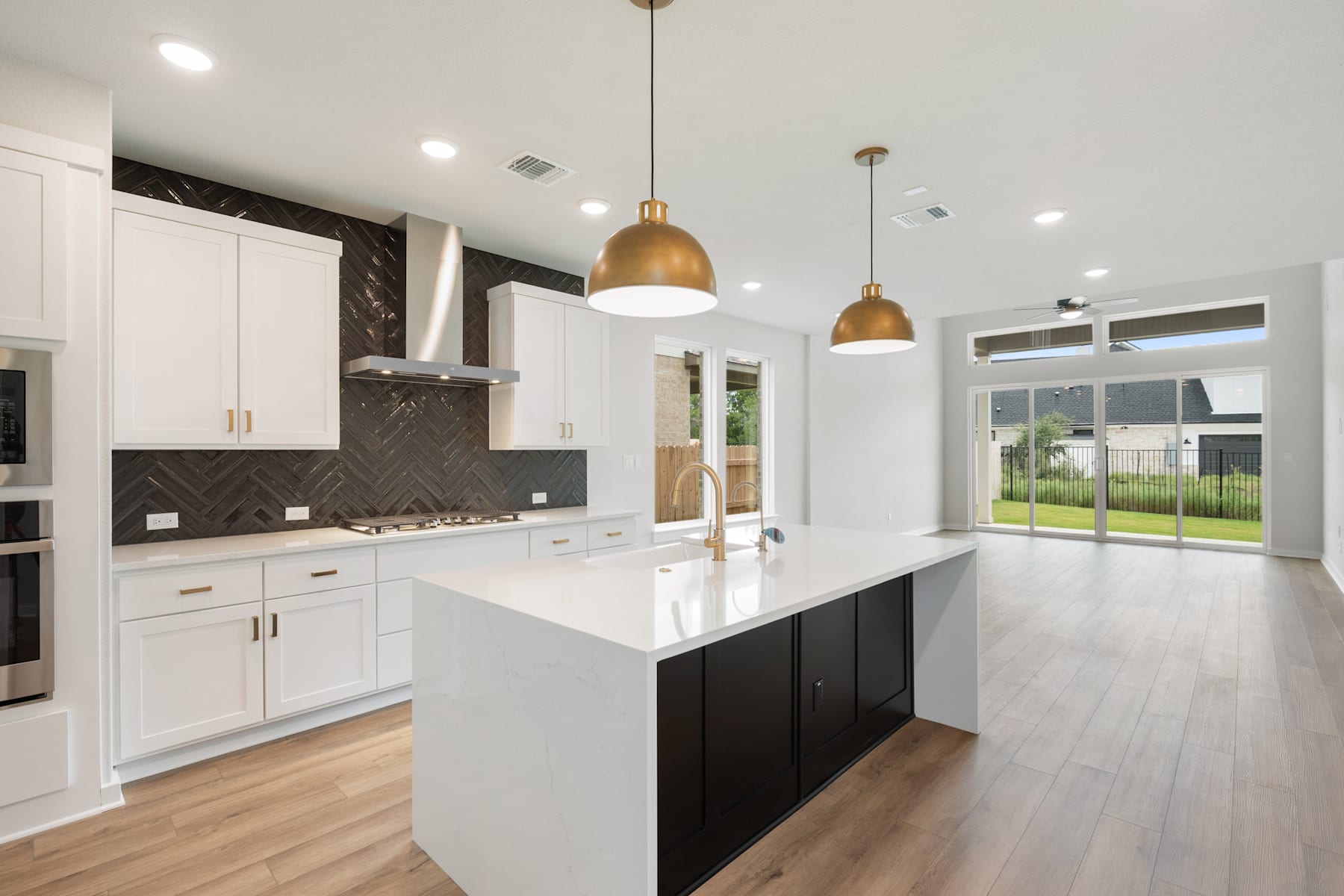 A modern, open-concept kitchen with white cabinets, a large island, and pendant lighting, set against a backdrop of hardwood floors and a sliding glass door leading to an outdoor area.