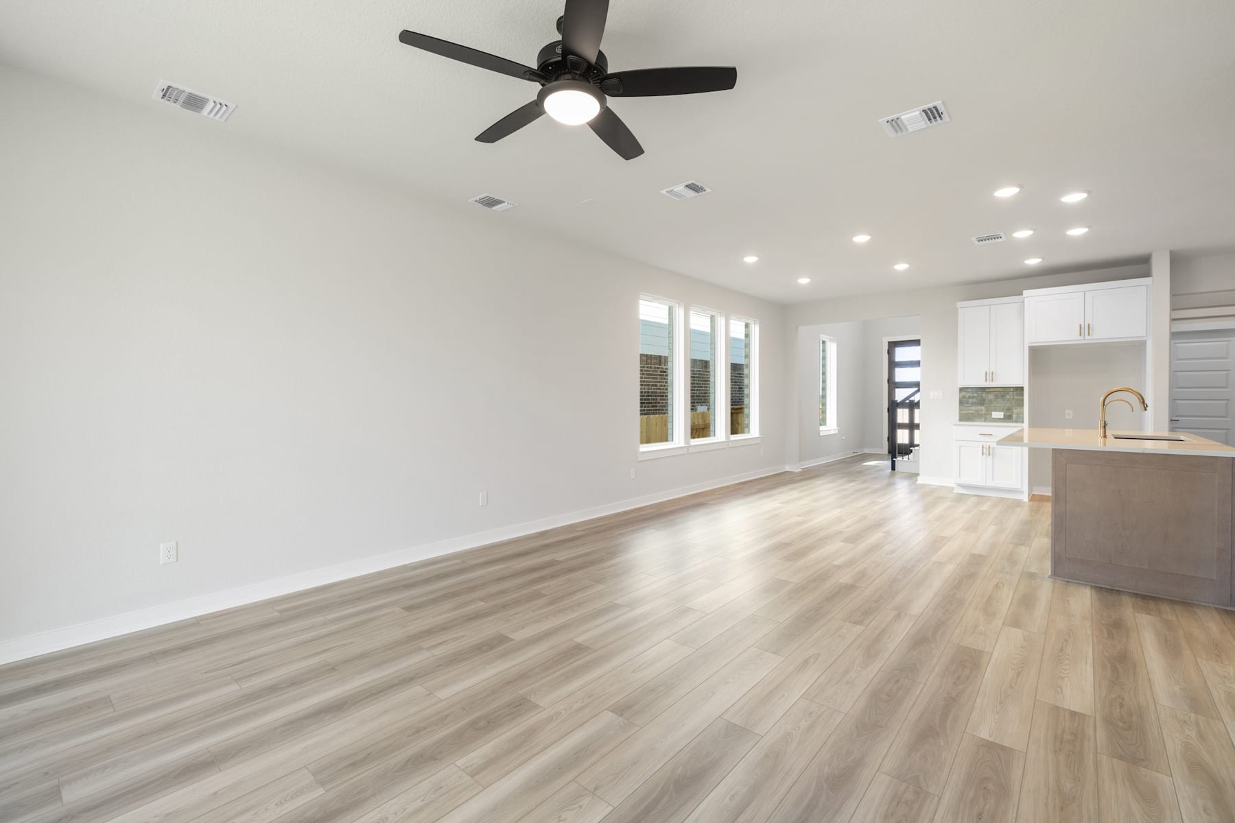 A spacious, well-lit room with a ceiling fan, hardwood floors, and a kitchen visible in the background.