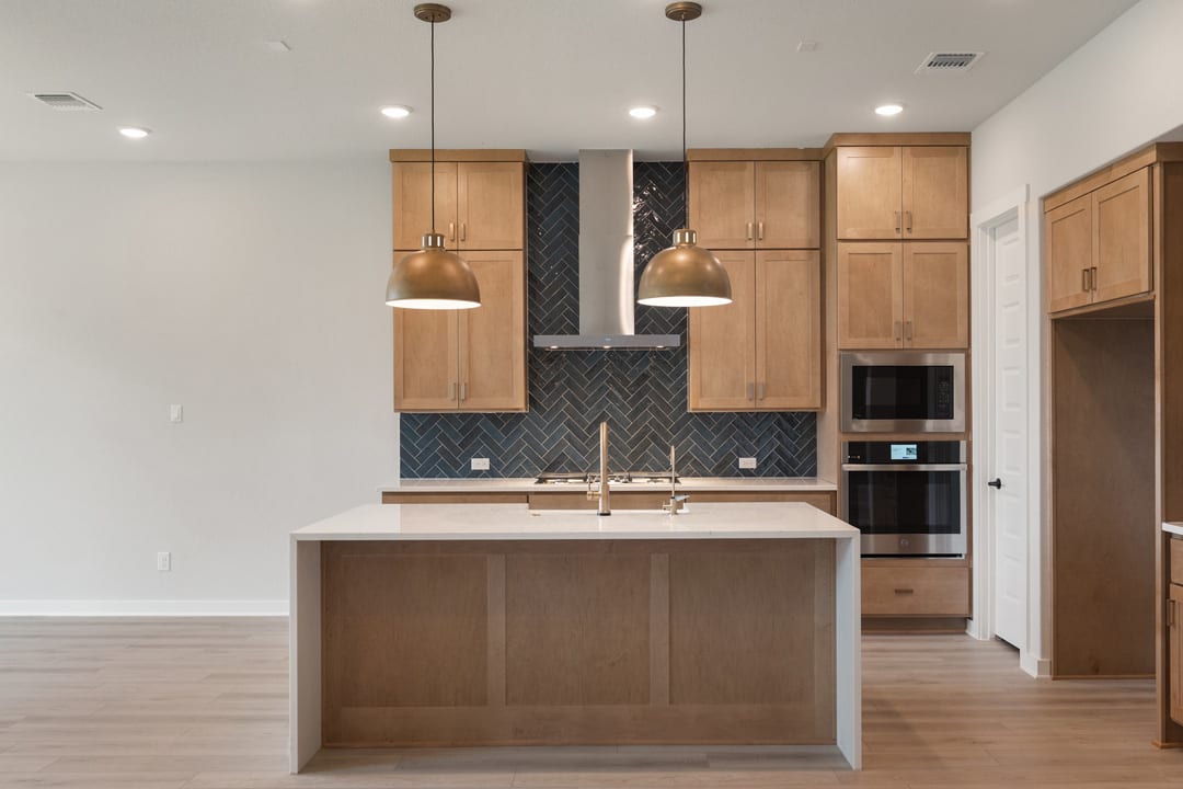 A modern kitchen with light wood cabinets, a central island, and pendant lighting fixtures, set against a backdrop of white walls and hardwood floors.