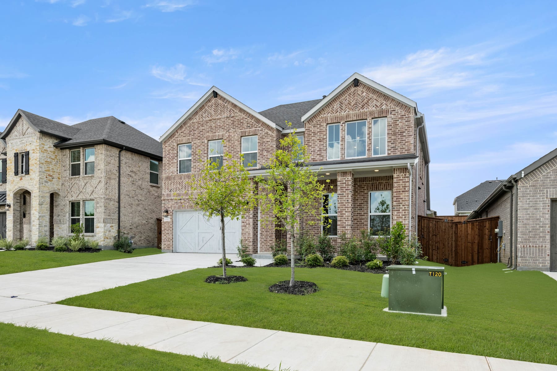 A two-story brick and stone residential house with a well-manicured lawn, surrounded by other similar houses in a suburban neighborhood under a clear blue sky.