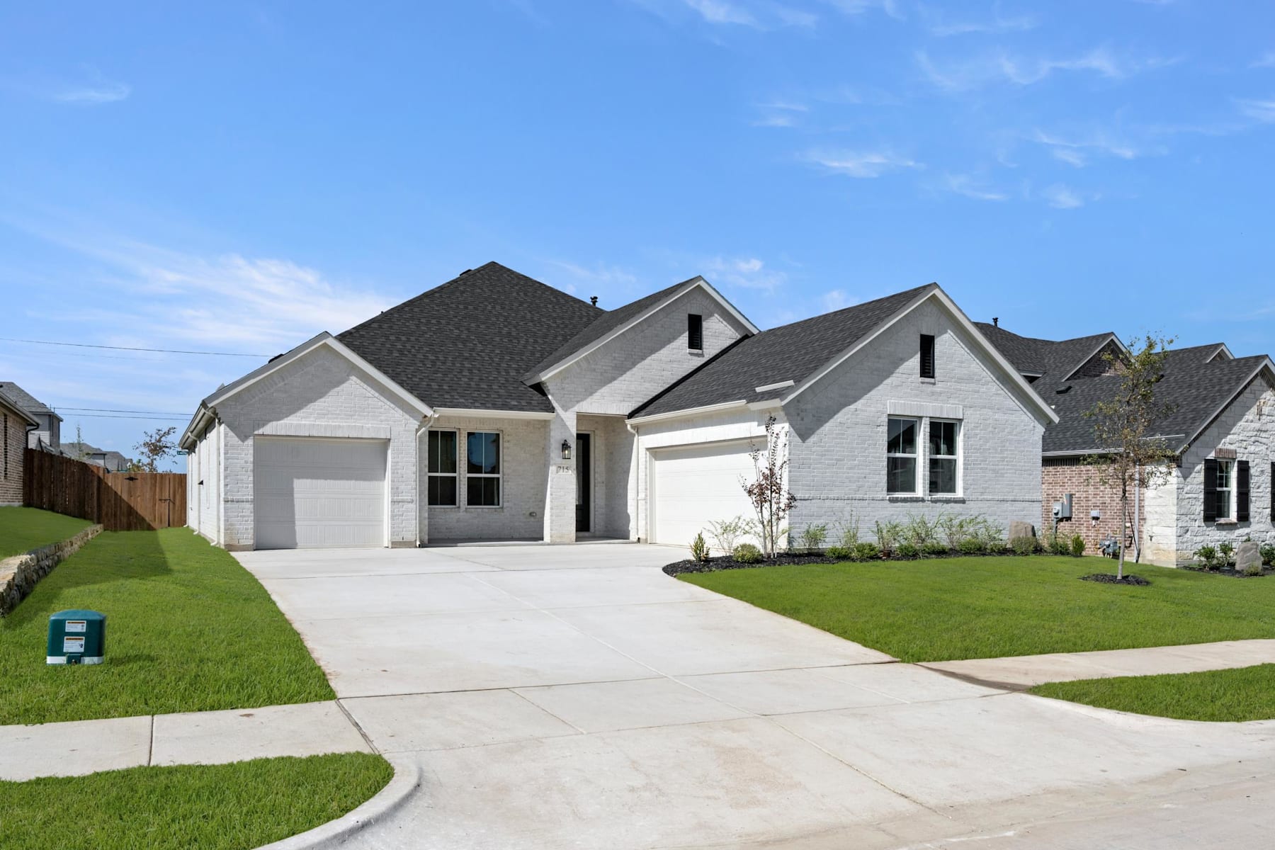 A newly constructed two-story house with a gray exterior, a black roof, and a well-manicured lawn in the foreground, set against a clear blue sky with scattered clouds.