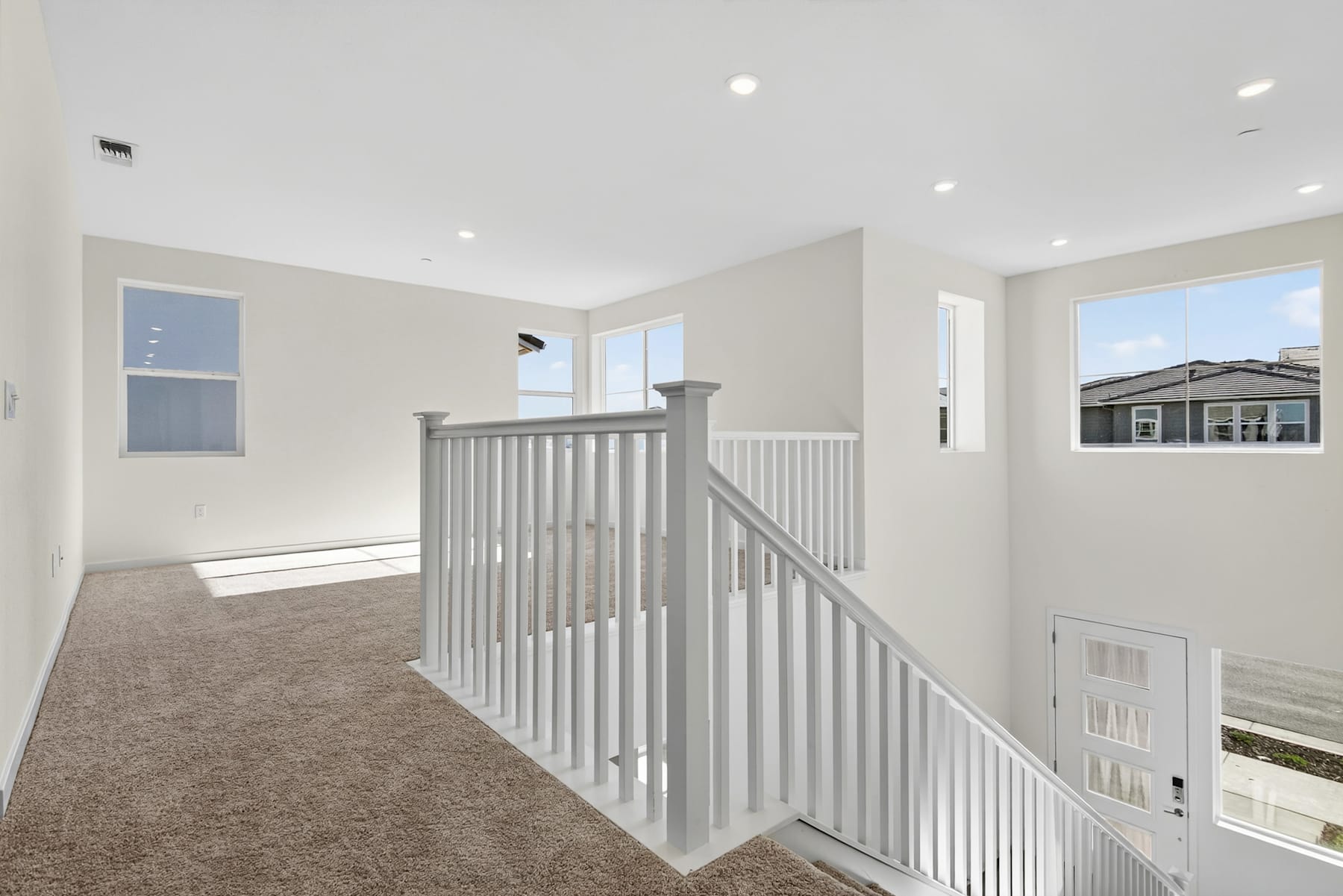 A bright and spacious hallway with a white staircase railing leading up to a second floor, surrounded by neutral-toned walls and carpeted floors.