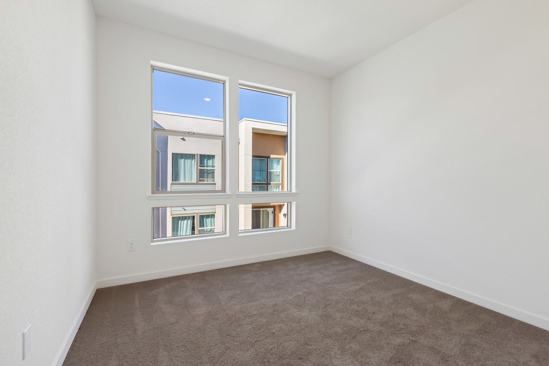 A bright, empty room with large windows overlooking a residential building in the background.