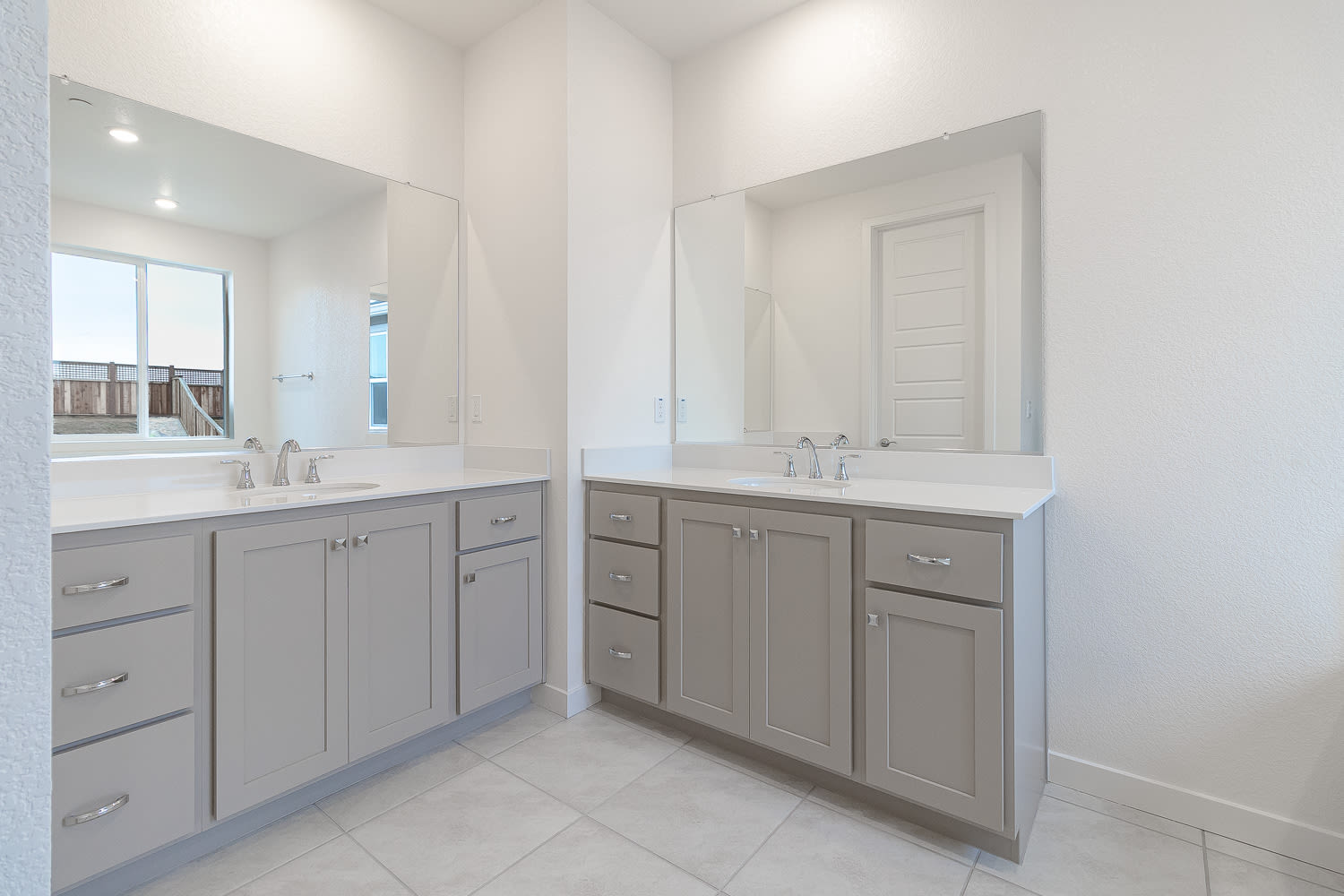 A modern, well-lit bathroom with gray cabinets, white countertops, and tiled floors, featuring two sinks and mirrors.