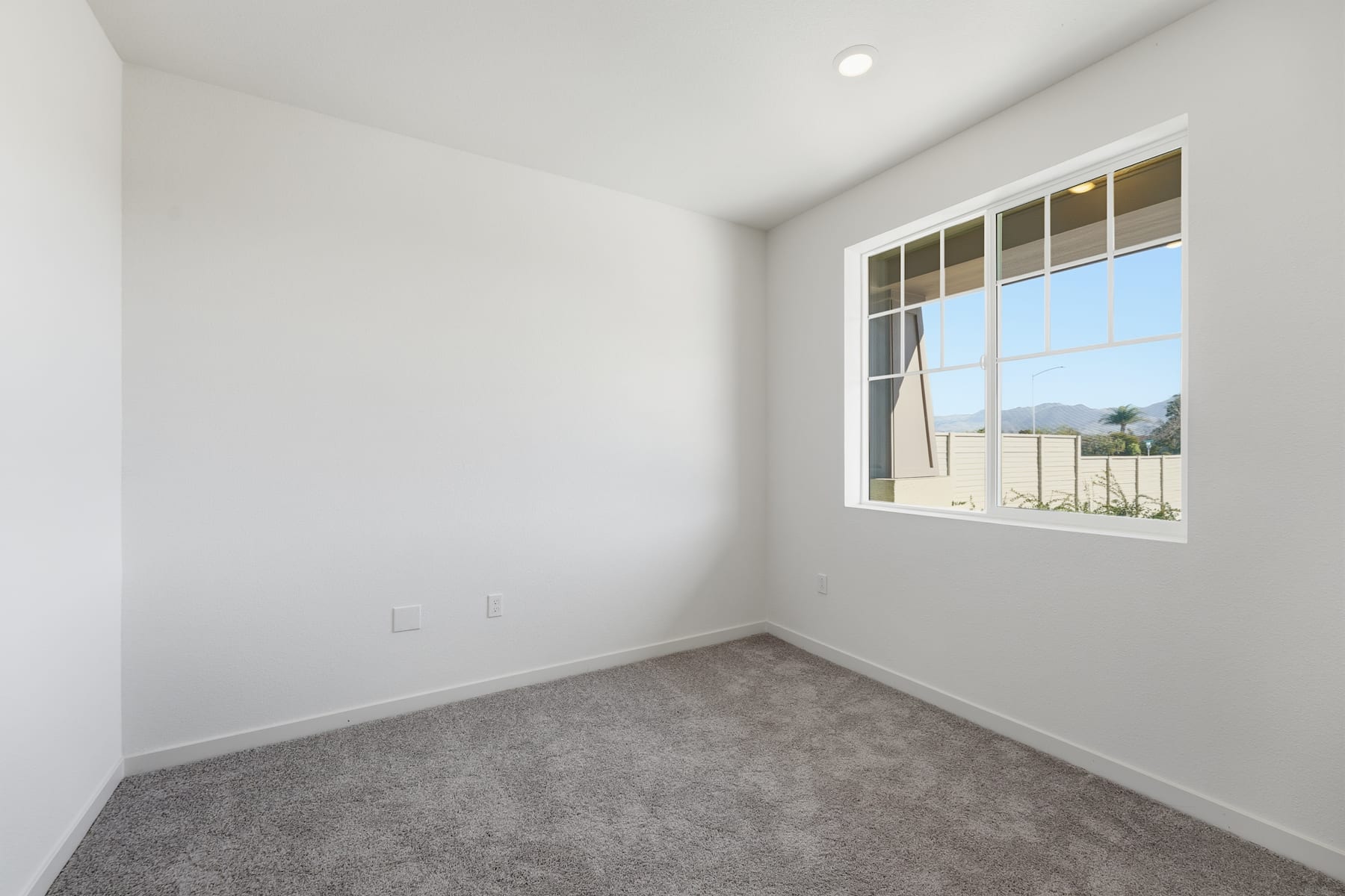 A bright, empty room with a large window overlooking an outdoor area, featuring a carpeted floor and plain white walls.