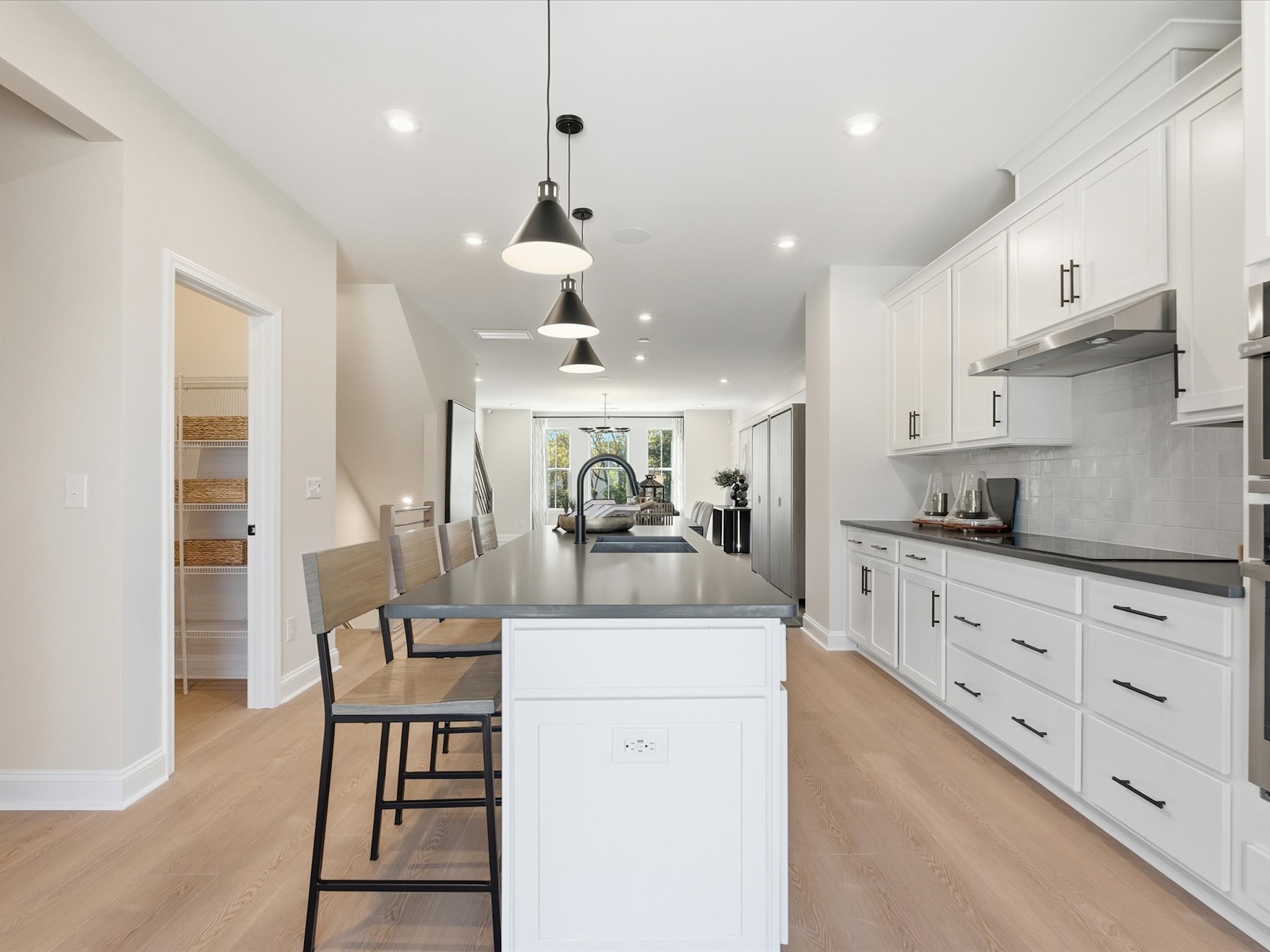 A modern, open-concept kitchen with white cabinets, a large island, and pendant lighting, leading into a hallway with a view of the outdoors.