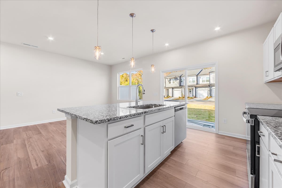 A modern, well-lit kitchen with gray cabinets, a granite countertop, and pendant lights, set against a backdrop of large windows overlooking an outdoor area.
