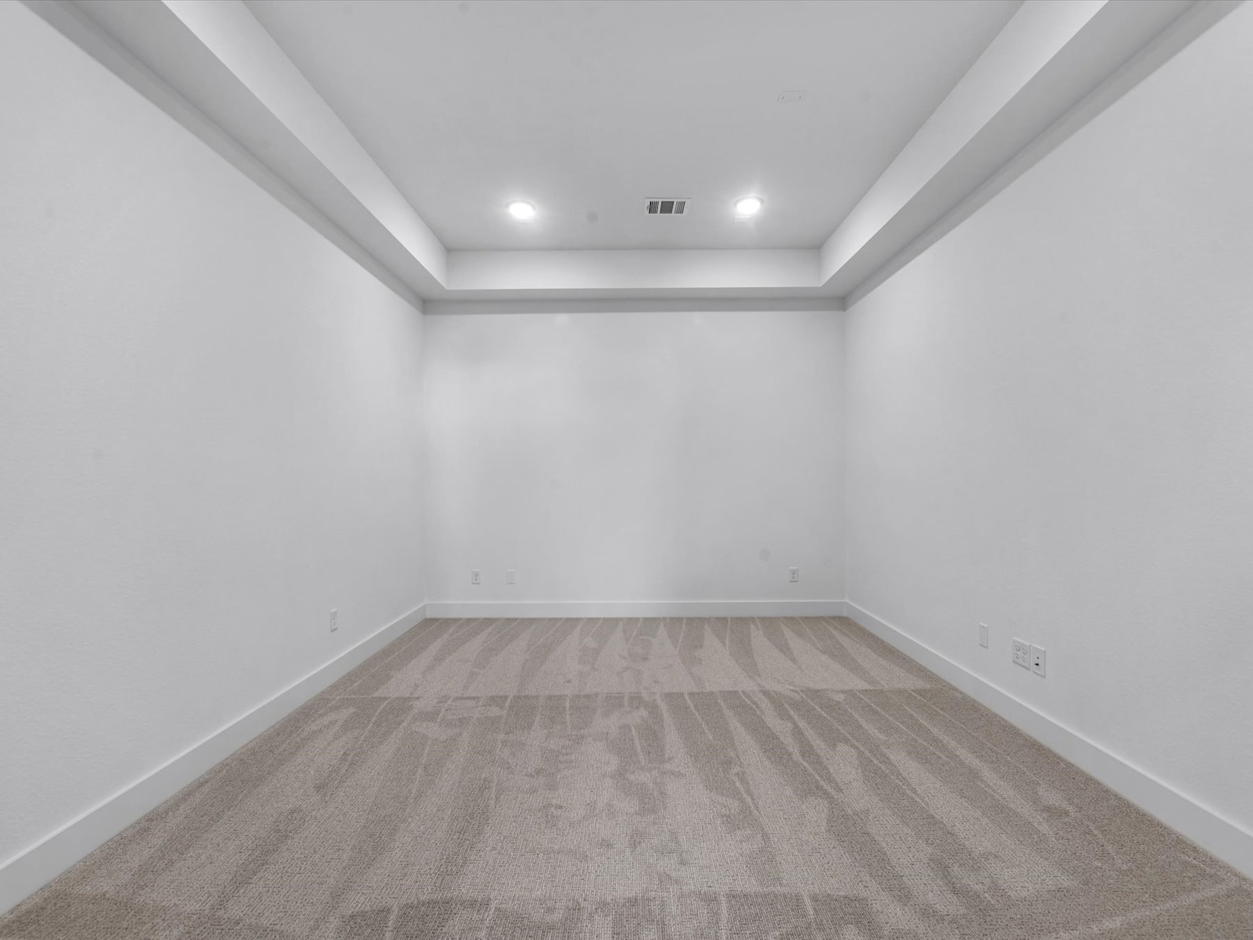 A spacious, empty room with white walls and a wooden floor, illuminated by recessed lighting.