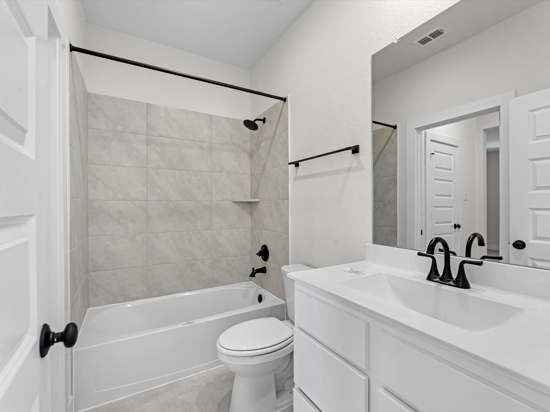 A modern, minimalist bathroom with a white tiled shower, a white vanity with a sink, and a toilet. The room has a clean, sleek aesthetic with black hardware accents.