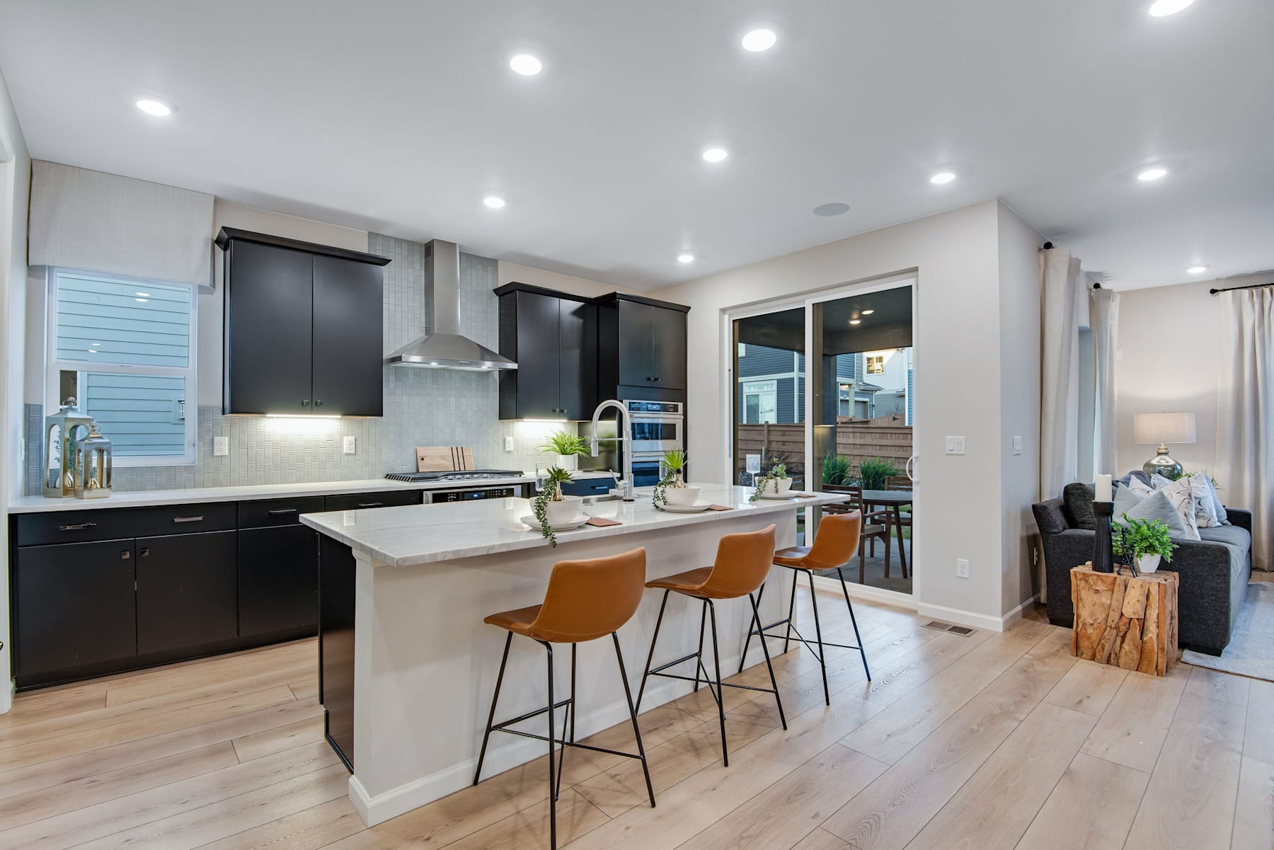 A modern, open-concept kitchen with sleek black cabinets, a white countertop, and bar stools, set against a backdrop of hardwood floors and recessed lighting.