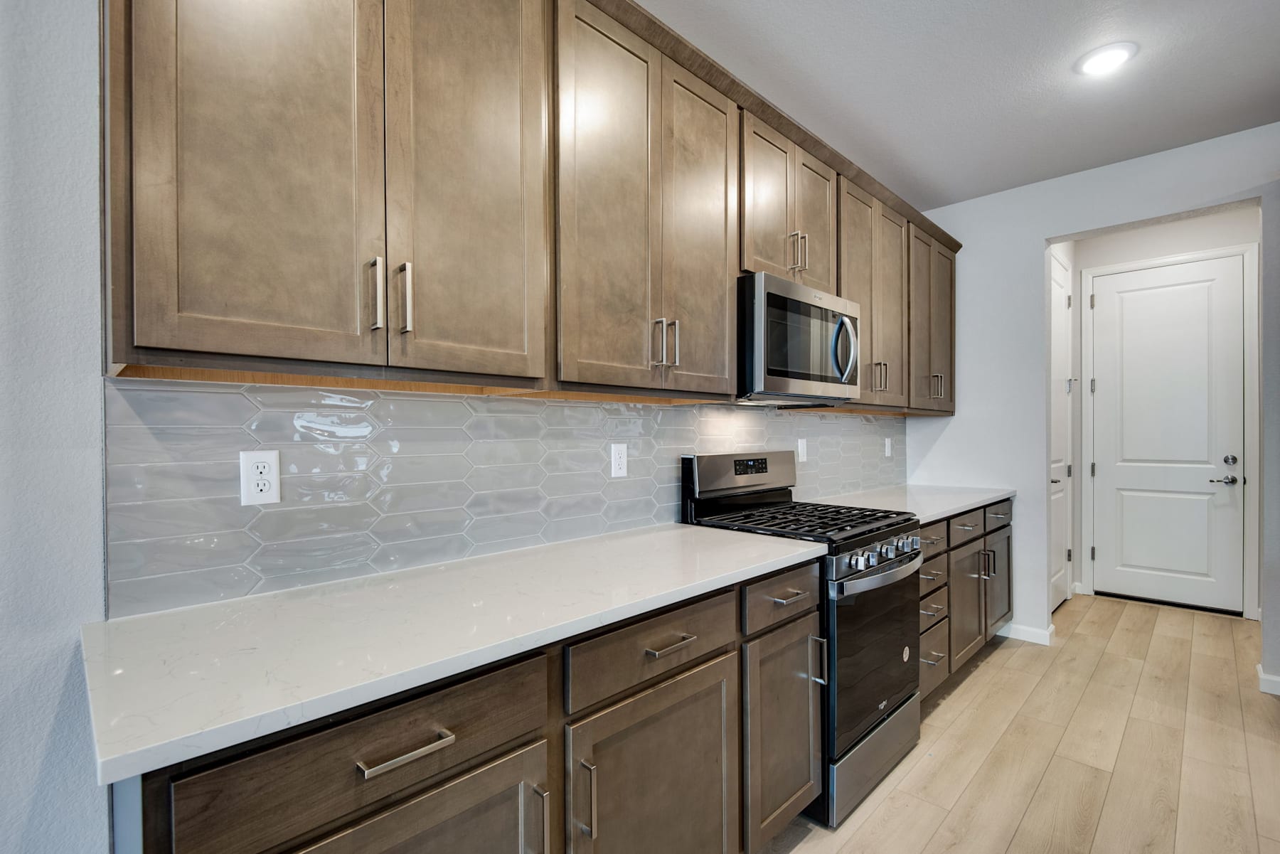 A modern kitchen with wooden cabinets, a white countertop, and stainless steel appliances, set against a tiled backsplash and hardwood flooring.