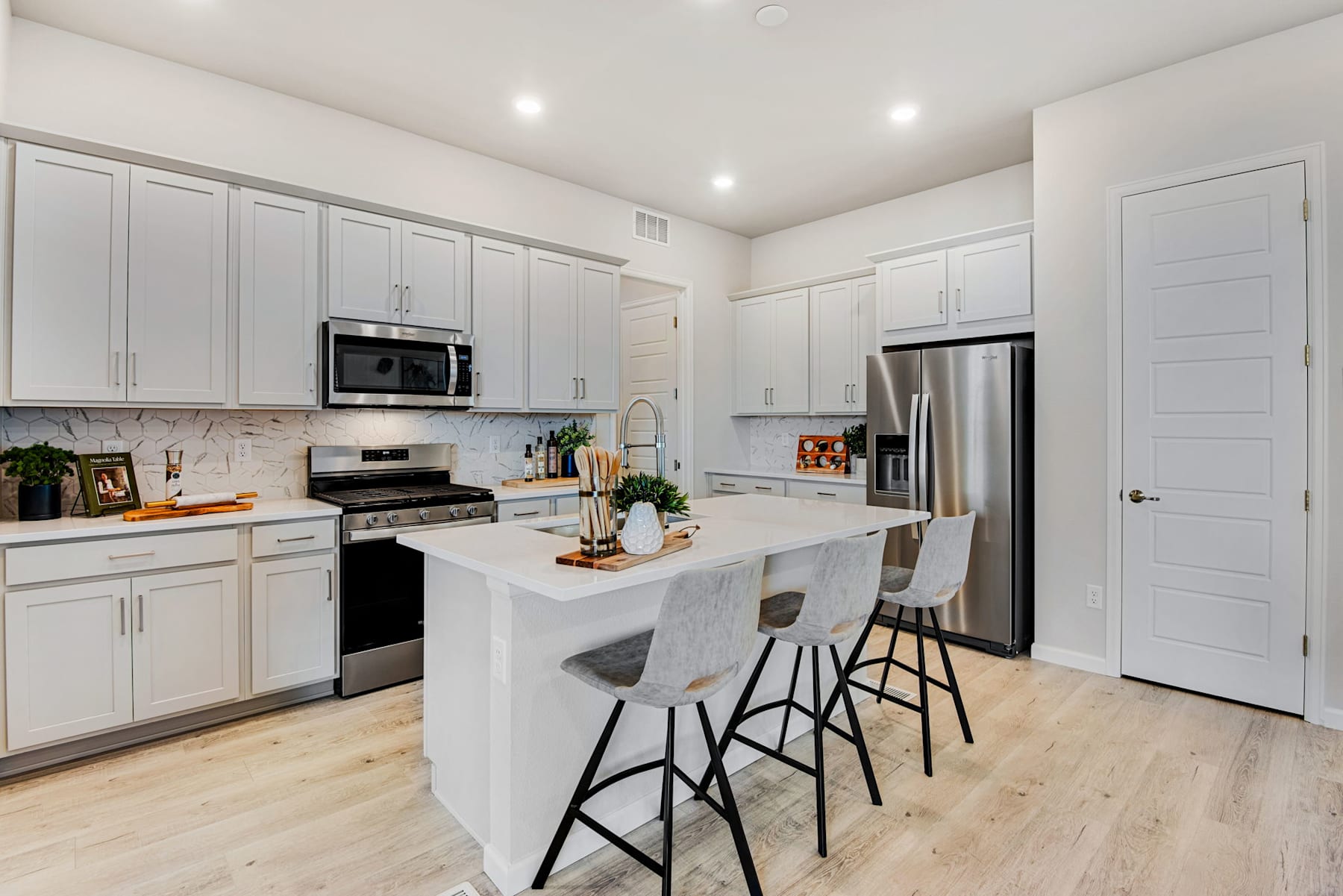 A modern, well-lit kitchen with white cabinets, stainless steel appliances, and a central island with bar stools, set against a backdrop of hardwood floors.